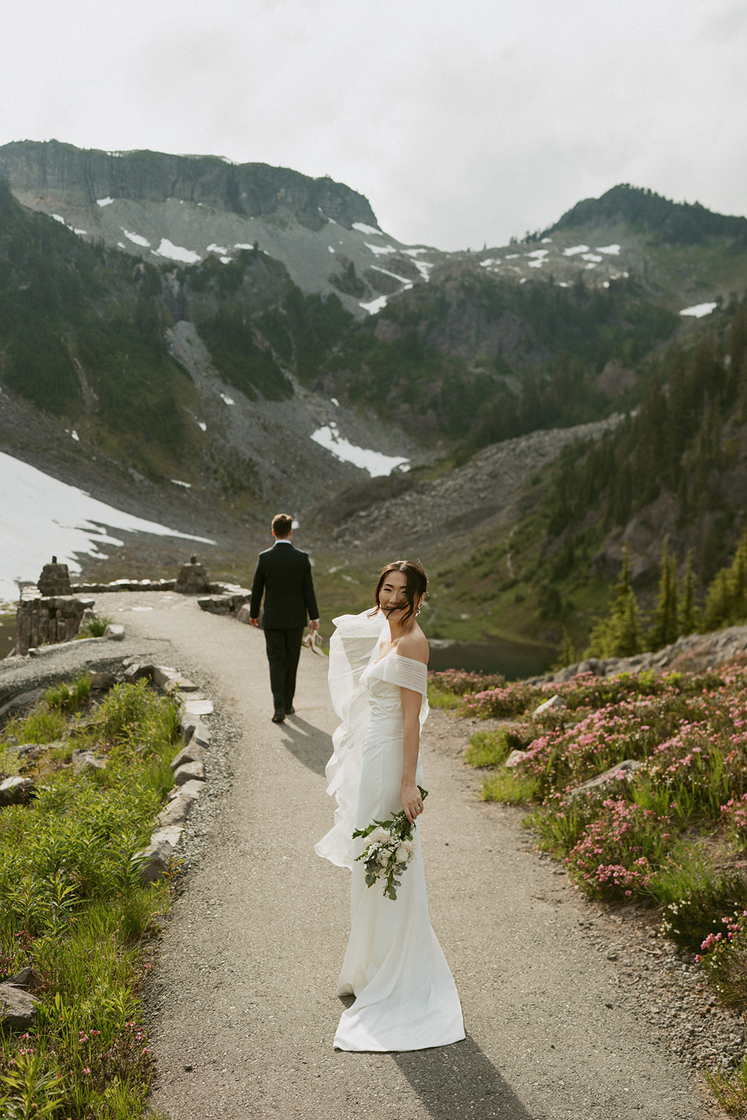 Bride glances over her shoulder on a mountain trail, bouquet in hand, with alpine ridges in the distance.