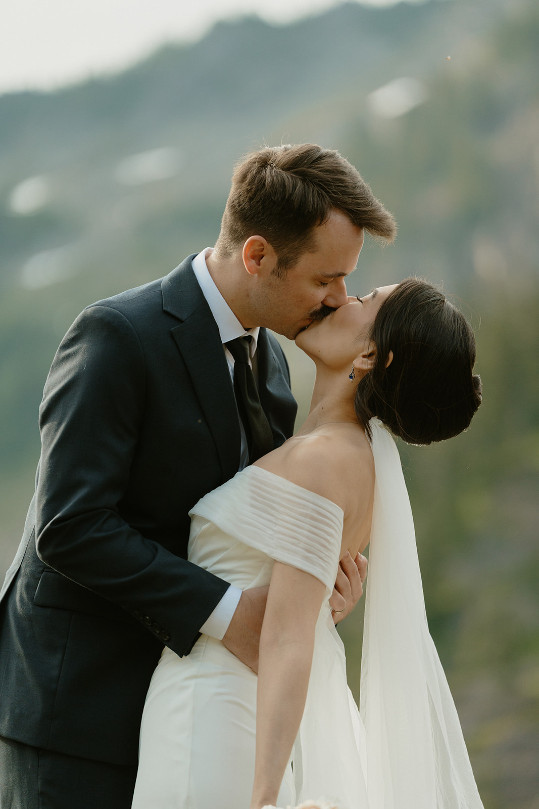 Groom leans in for a kiss with his bride, surrounded by forested mountain slopes.