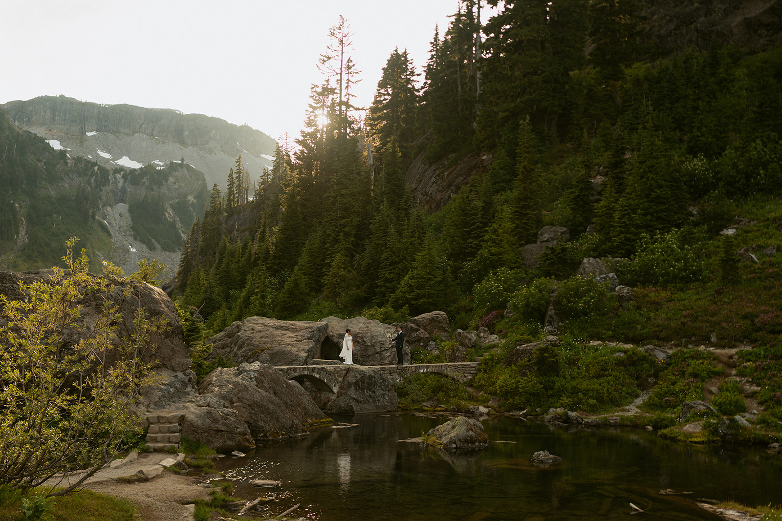 Couple crosses a mountain stream at sunset while eloping in Washington’s rugged alpine landscape.
