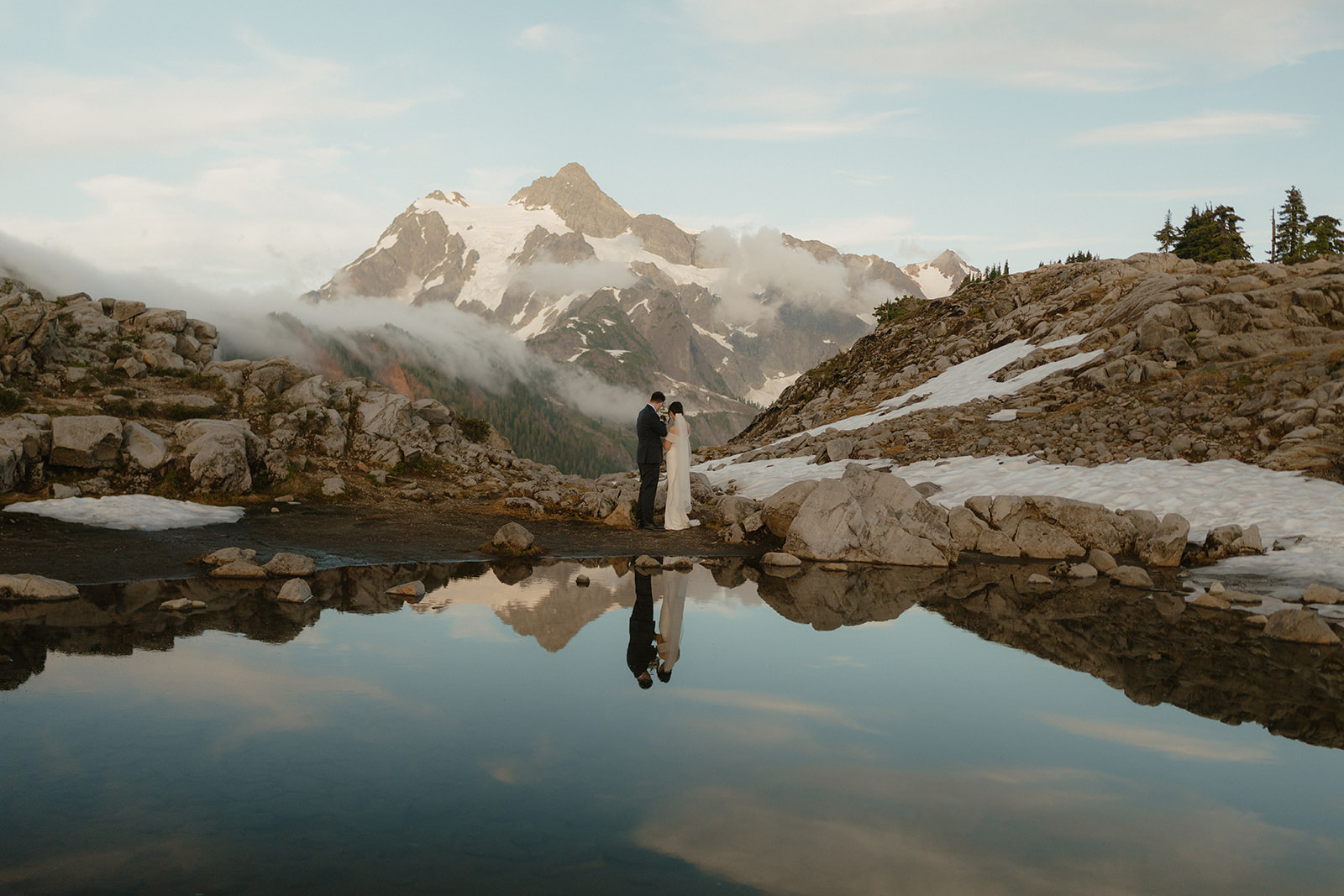 Couple stands together, mirrored in an alpine lake beneath Mount Shuksan, a highlight of eloping in Washington.