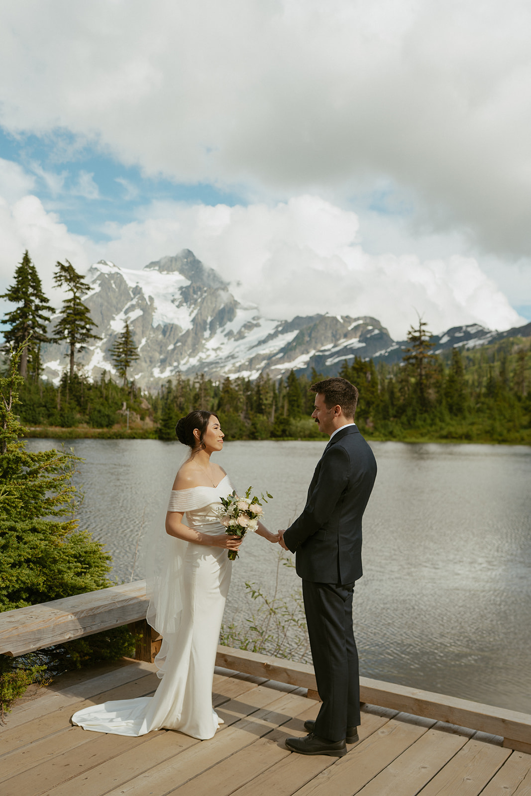 Eloping in Washington, a couple stands together on a dock with mountain reflections behind them.