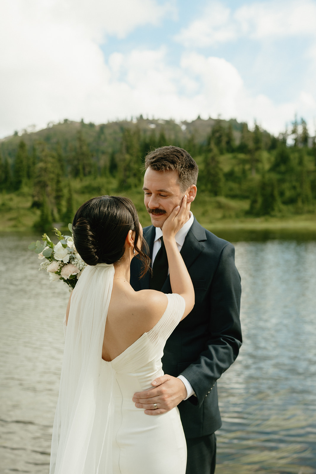 Bride gently touches her groom’s face during a quiet moment by the lake.