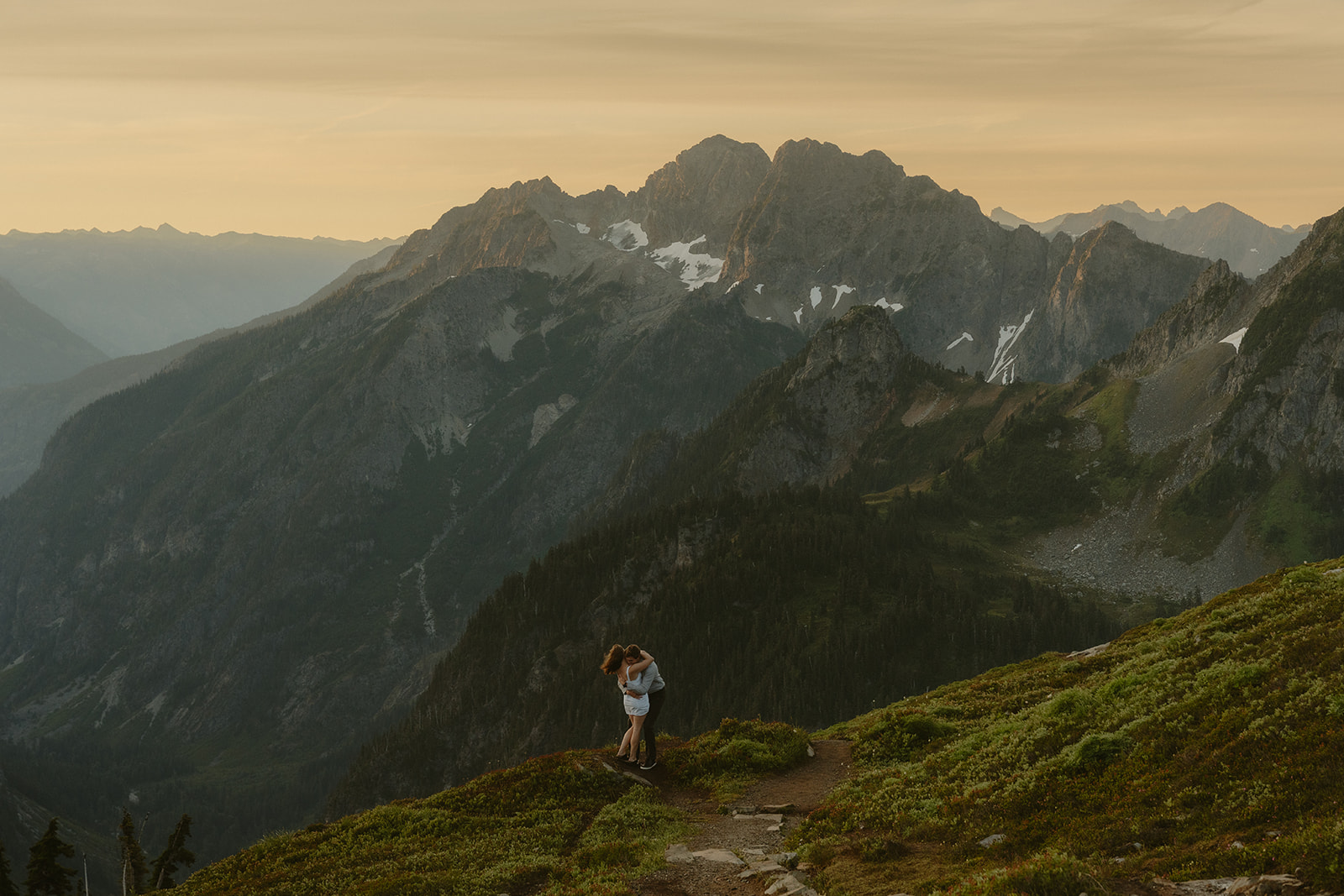 Couple embracing on a trail overlooking alpine ridges and a golden sunset—an intimate scene from their outdoor engagement photos in the Cascades.