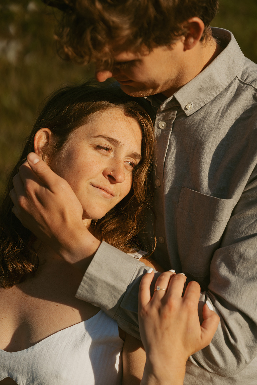 A tender moment as the man gently holds his partner’s face in golden light, her engagement ring catching the sun.