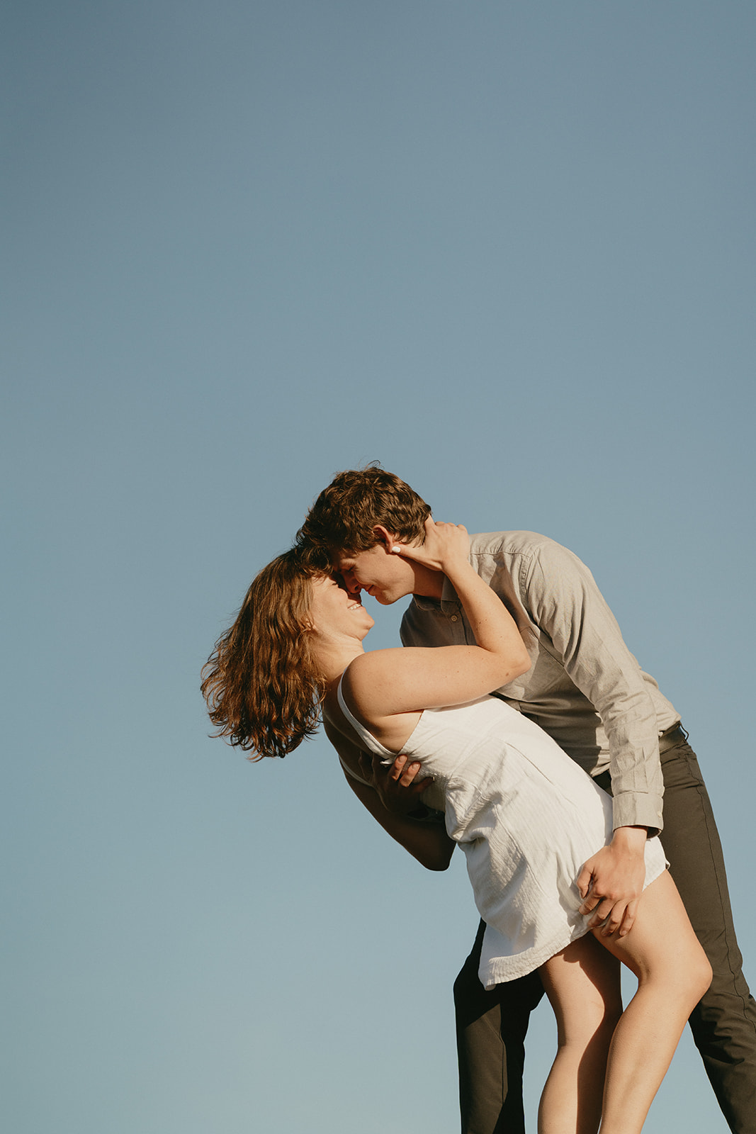 A man and woman share a kiss on a hilltop, surrounded by a scenic landscape under a clear blue sky.