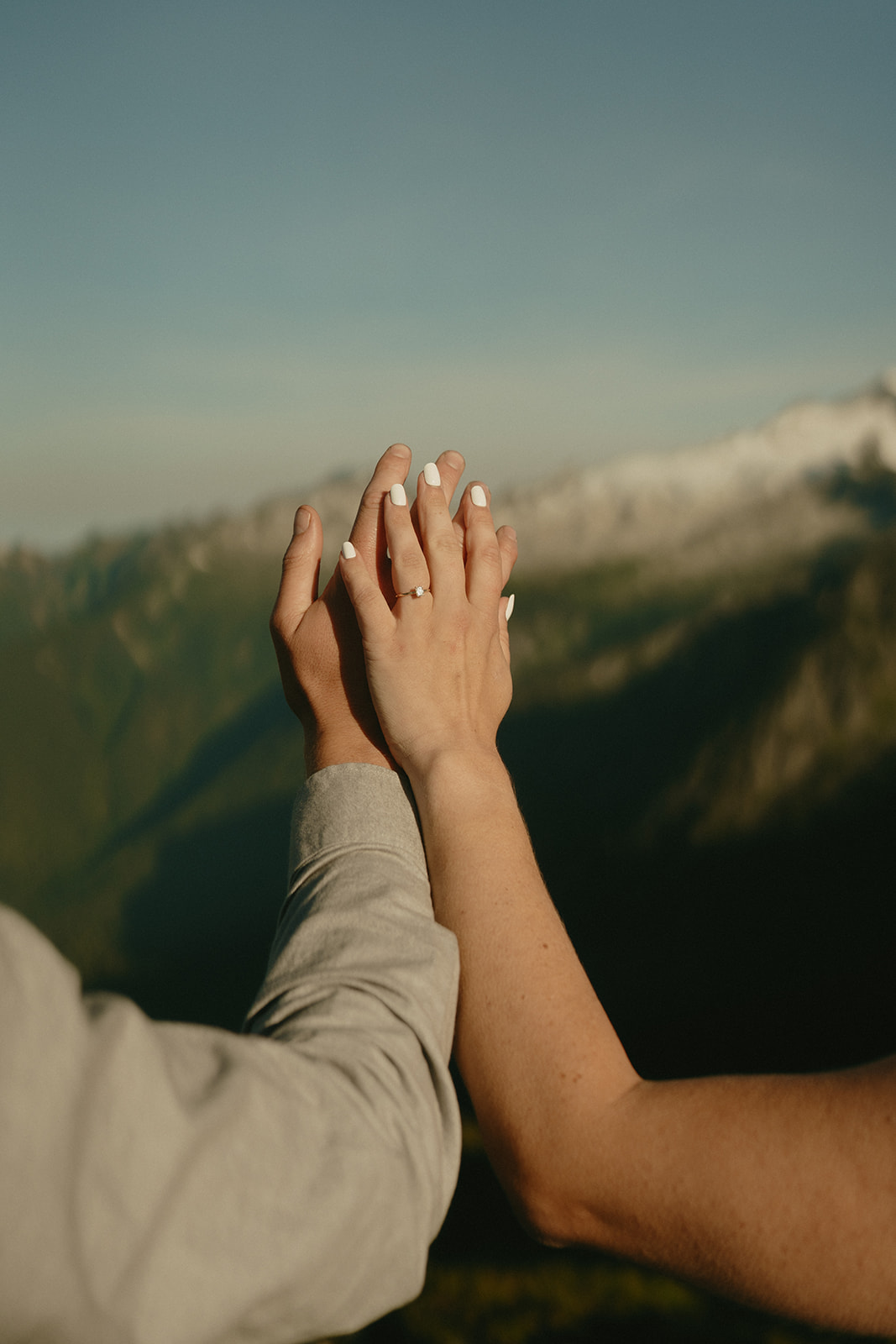 Hands reaching toward the sky with matching engagement rings, framed by a deep green valley and sunlit mountain peaks—symbolic and timeless outdoor engagement photo.