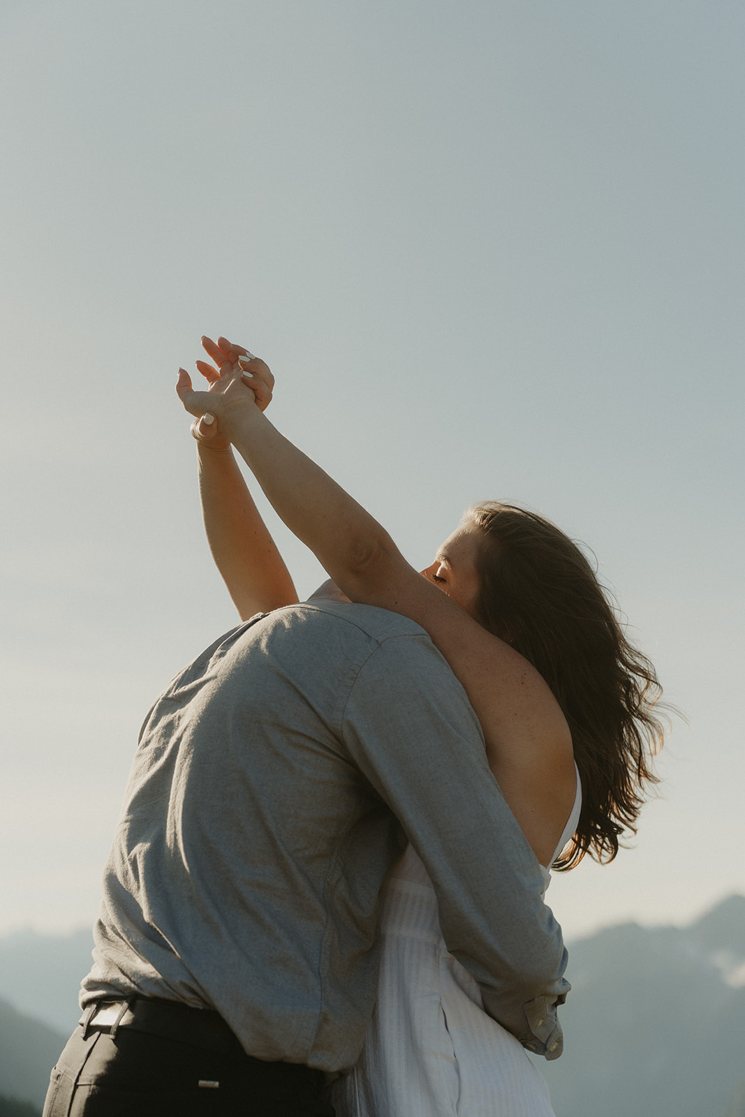 Couple embracing under a wide open sky, with her arms lifted in joy—an expressive and candid moment from their outdoor engagement photos in the North Cascades.