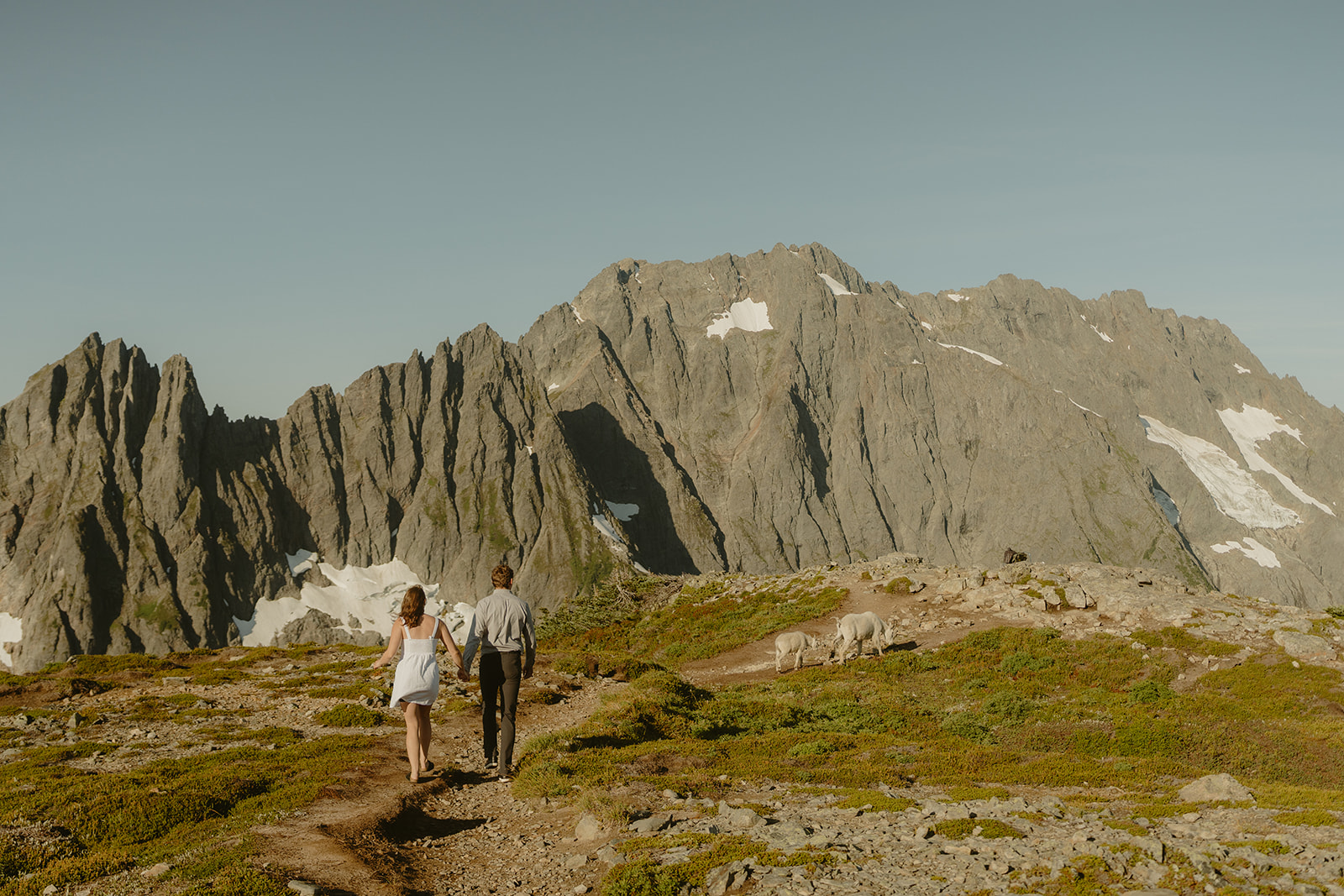 A couple walking hand in hand along a scenic mountain trail surrounded by lush greenery and towering peaks.
