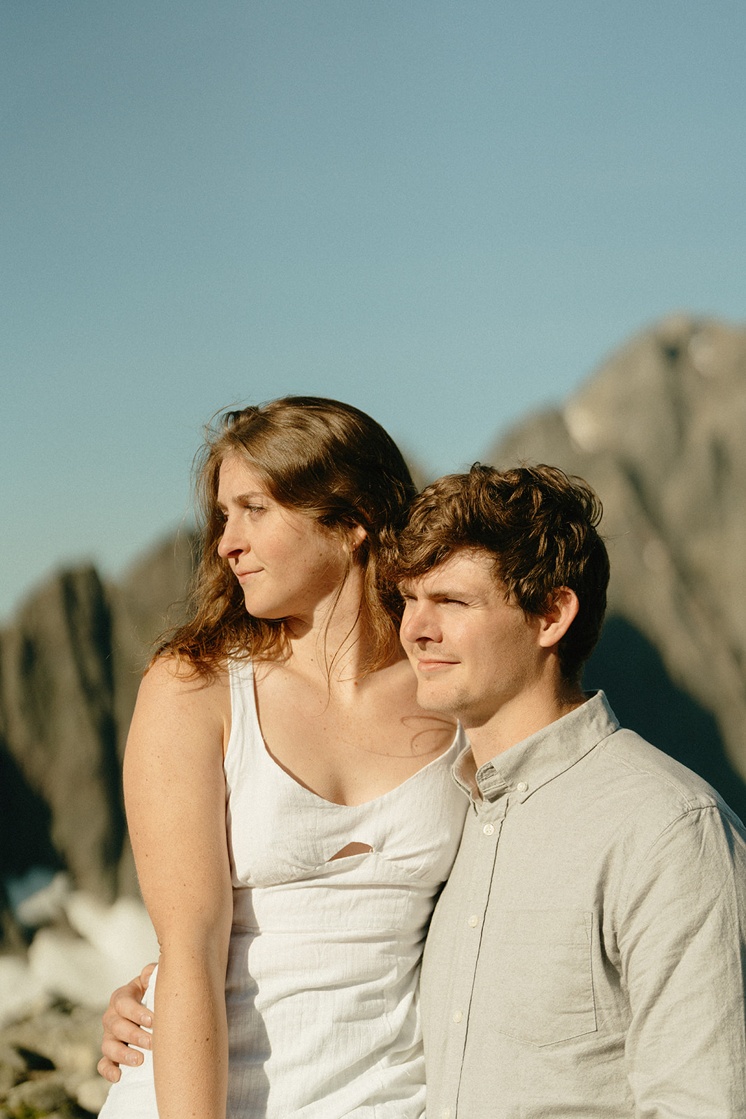 Couple gazes into the distance, wrapped in afternoon light with dramatic alpine cliffs rising behind them—part of their outdoor engagement photos in the mountains.