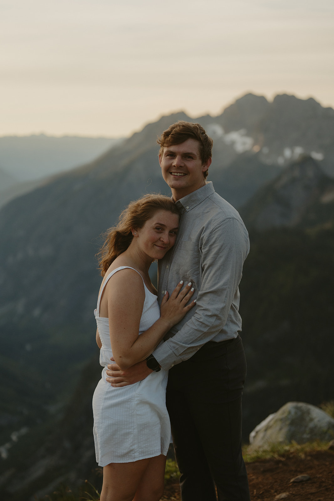 A couple embraces on a rocky mountain peak during their engagement session, surrounded by breathtaking scenic views.