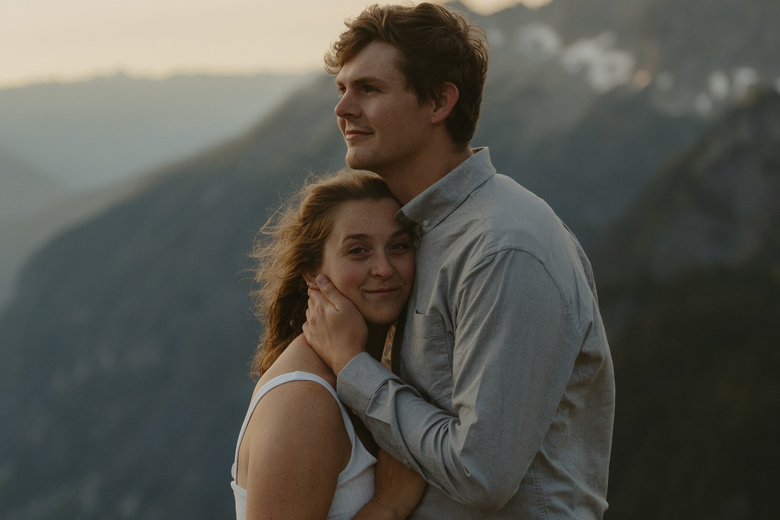 Woman nestles into her partner’s chest as he holds her gently, surrounded by hazy mountain layers at twilight.