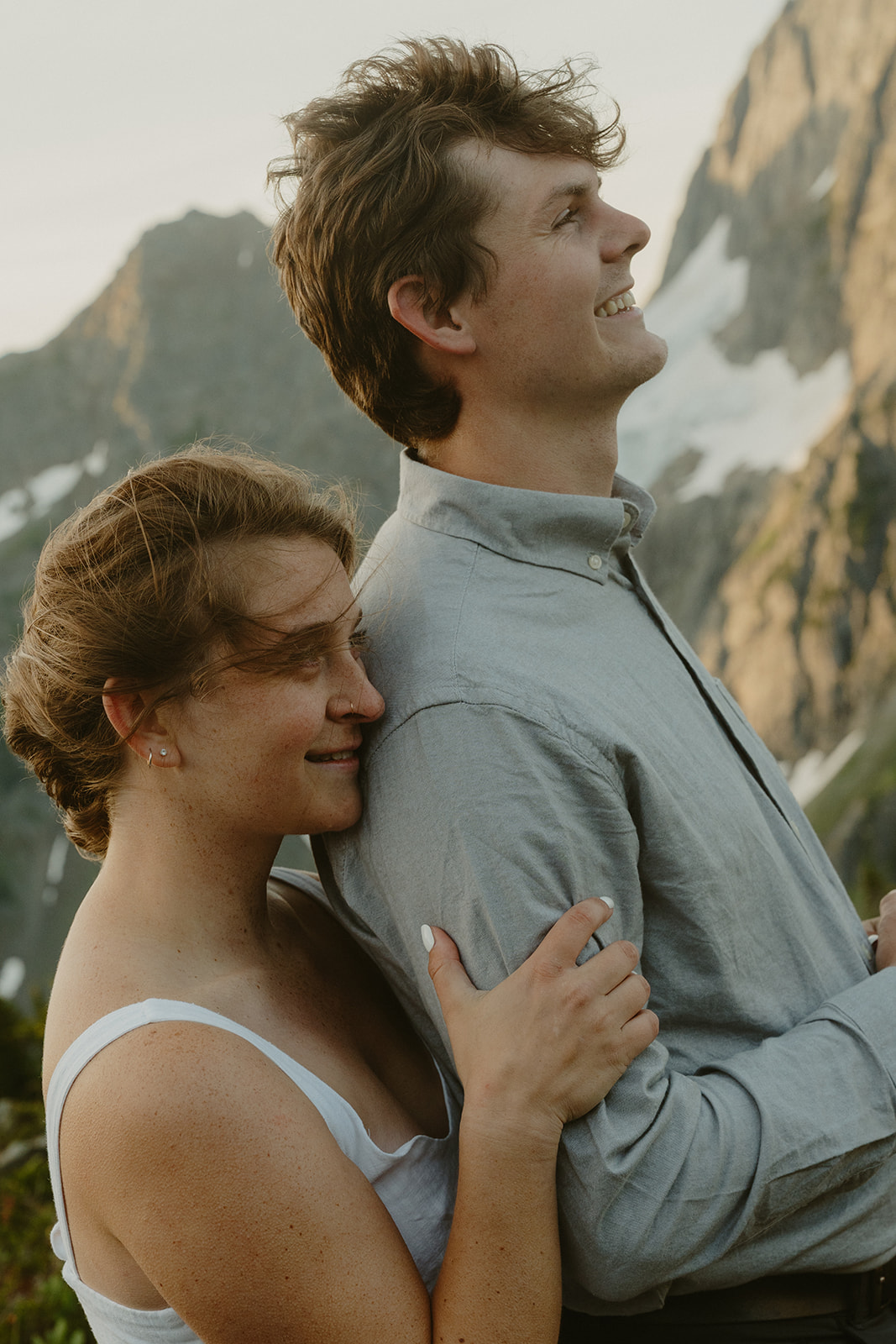 Woman holding man's arm from behind while man smiles looking at beautiful mountain range