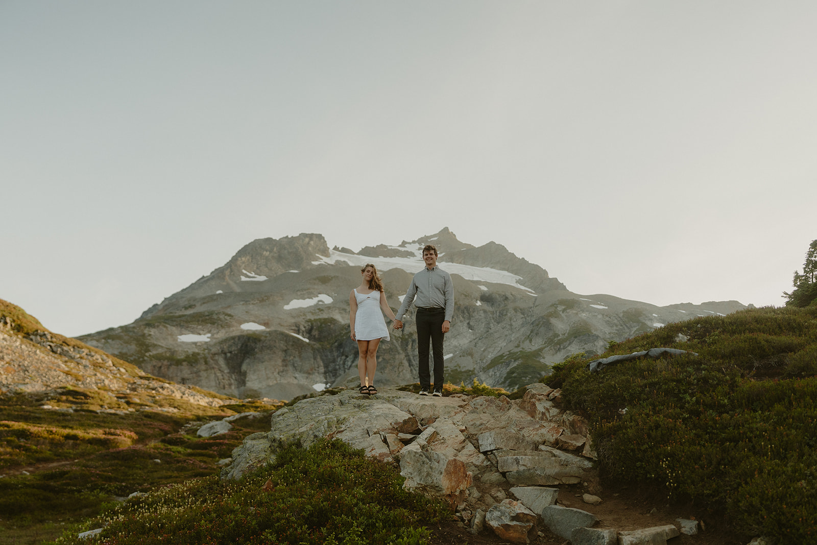 Couple standing hand-in-hand on a rocky slope, with snow-dusted mountain peaks rising behind them under a soft evening sky.