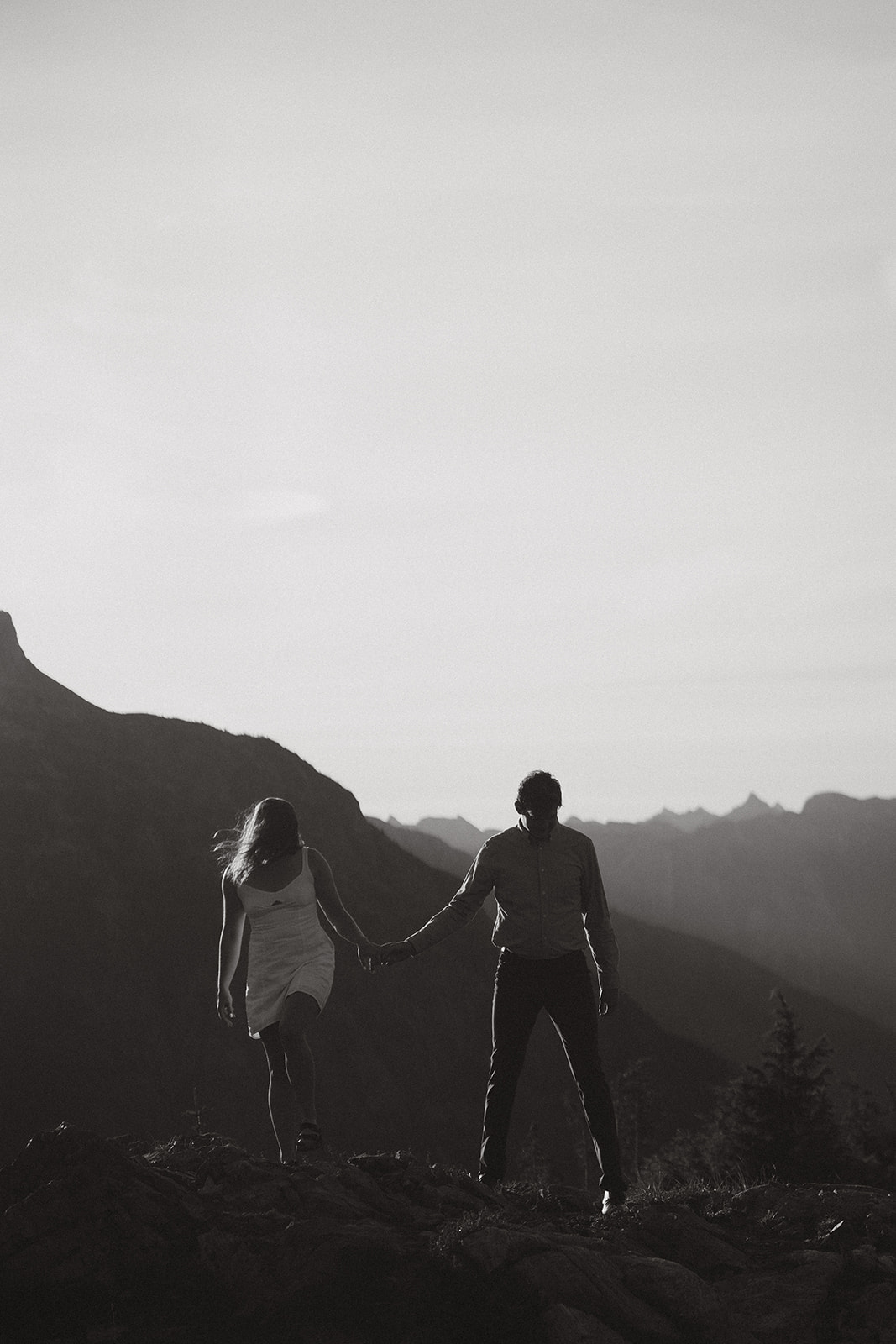 Silhouetted couple walking hand-in-hand across a ridgeline, backlit by soft evening light in the Pacific Northwest wilderness.