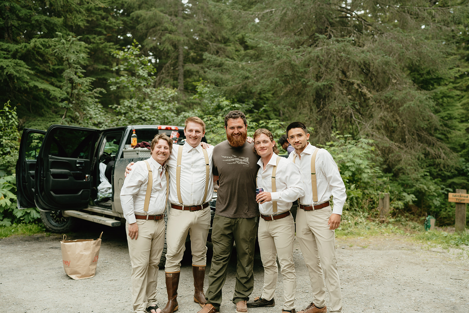 Groom stands with groomsmen before getting ready for his big day while groomsmen wear suspenders and khaki pants