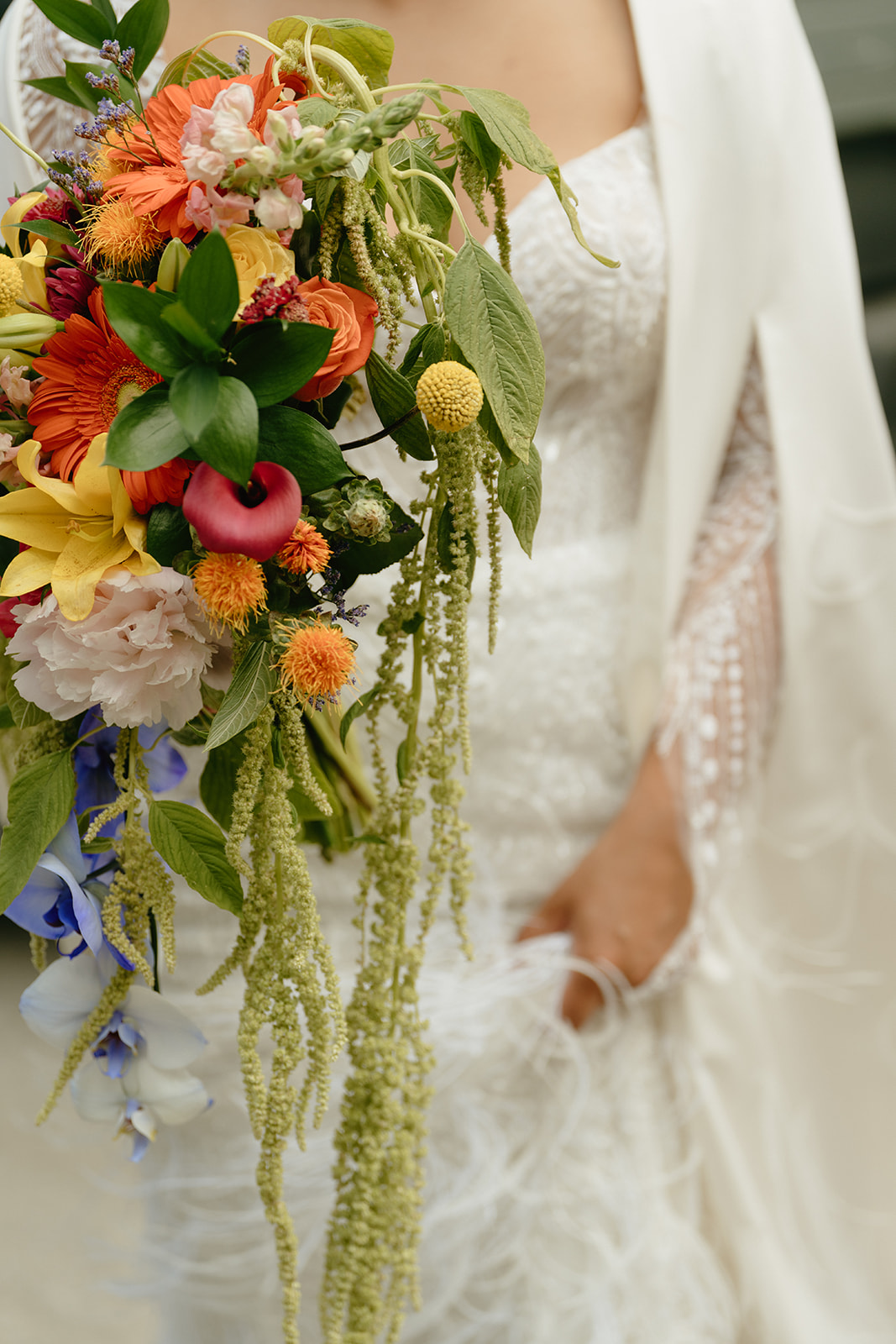 Close up of bride holding colorful bridal bouquet while lifting train to walk through forest.