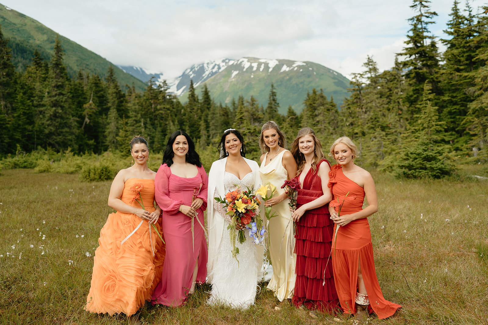 Bride stands with bridesmaids all wearing pink, orange, and yellow dresses mismatched.