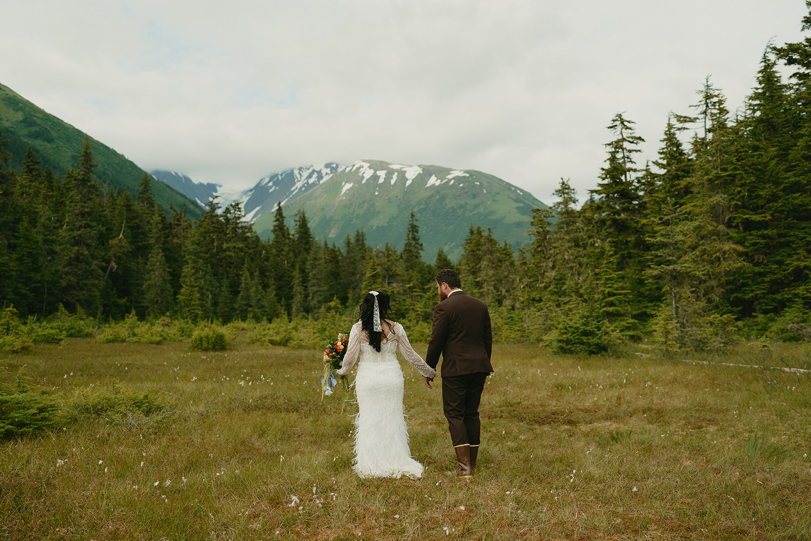 Wide shot of newly married couple walking hand in hand through grassy area with Alaska mountains in the distance