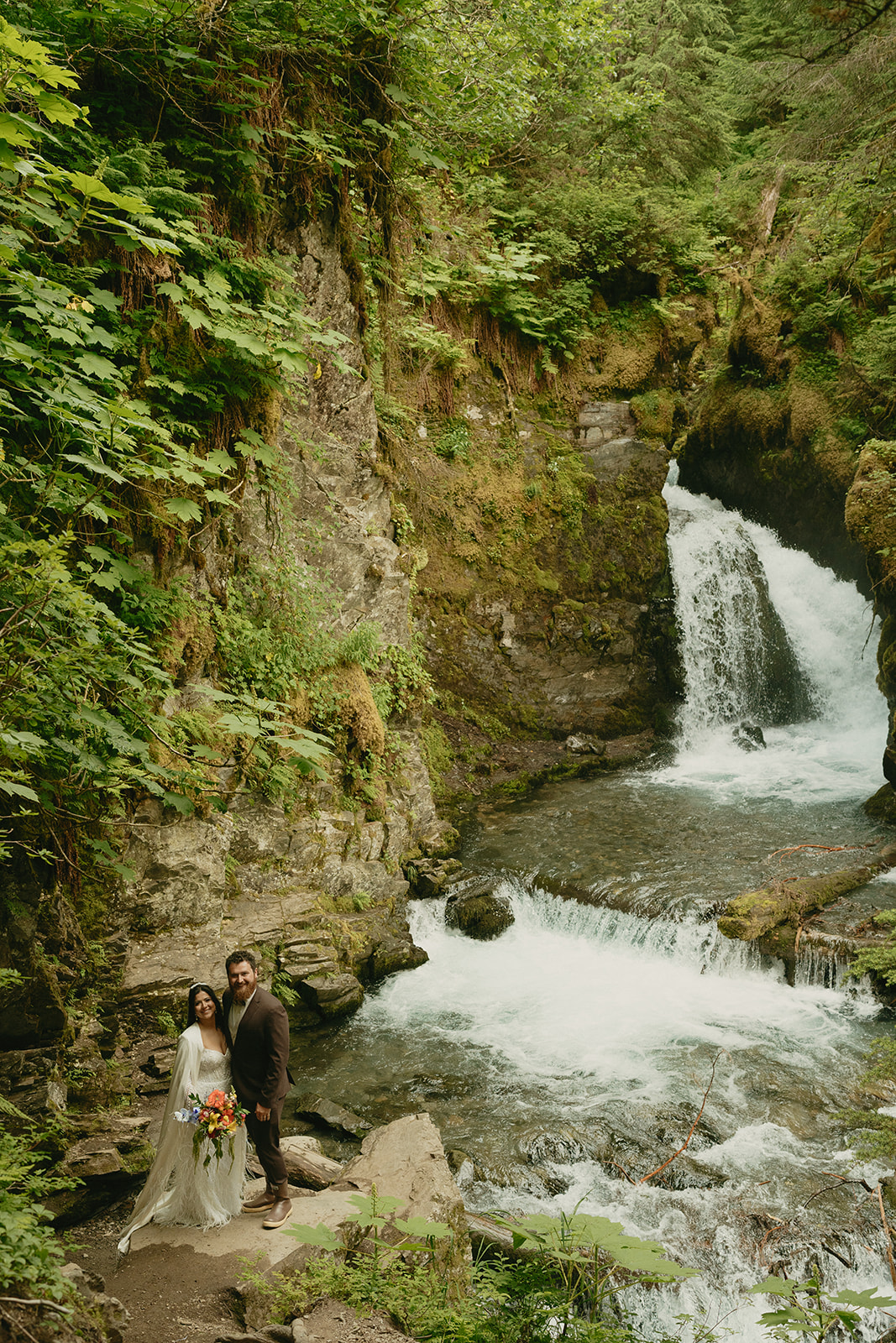 Couple stands beside waterfall embracing and smiling