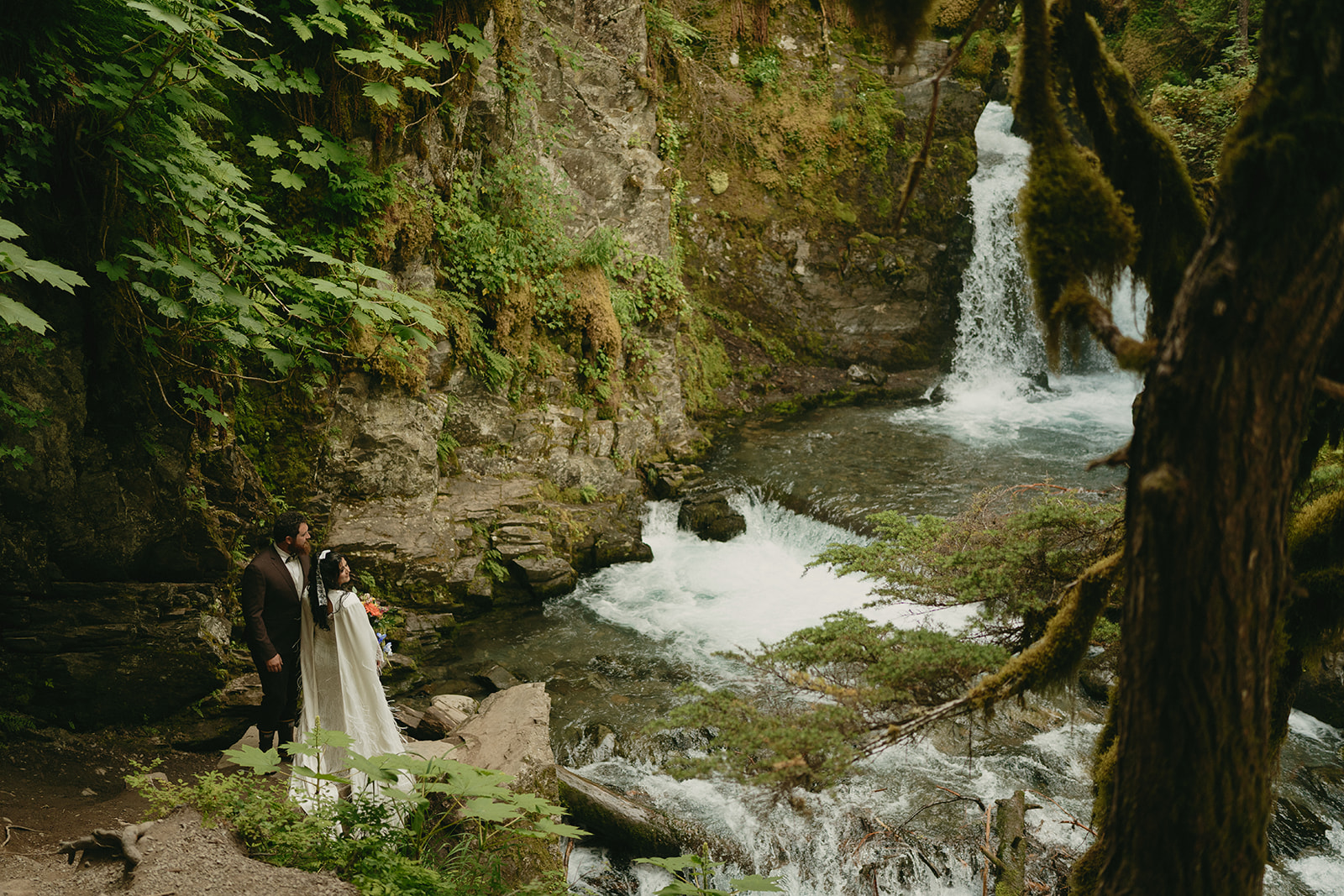 Man and woman stand together as newly married couple looking at the waterfall in the forested area they said their vows
