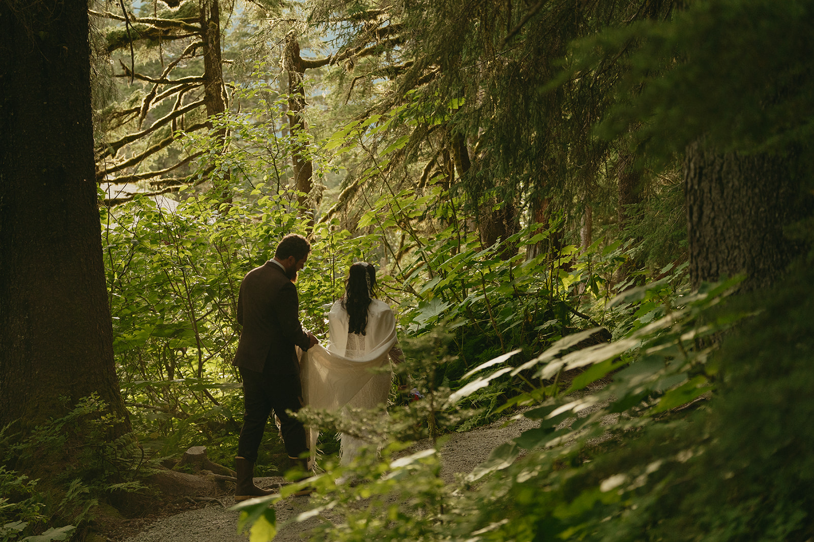 Bride and groom go for a private walk through forested area while groom holds the bride's cape after eloping to Alaska