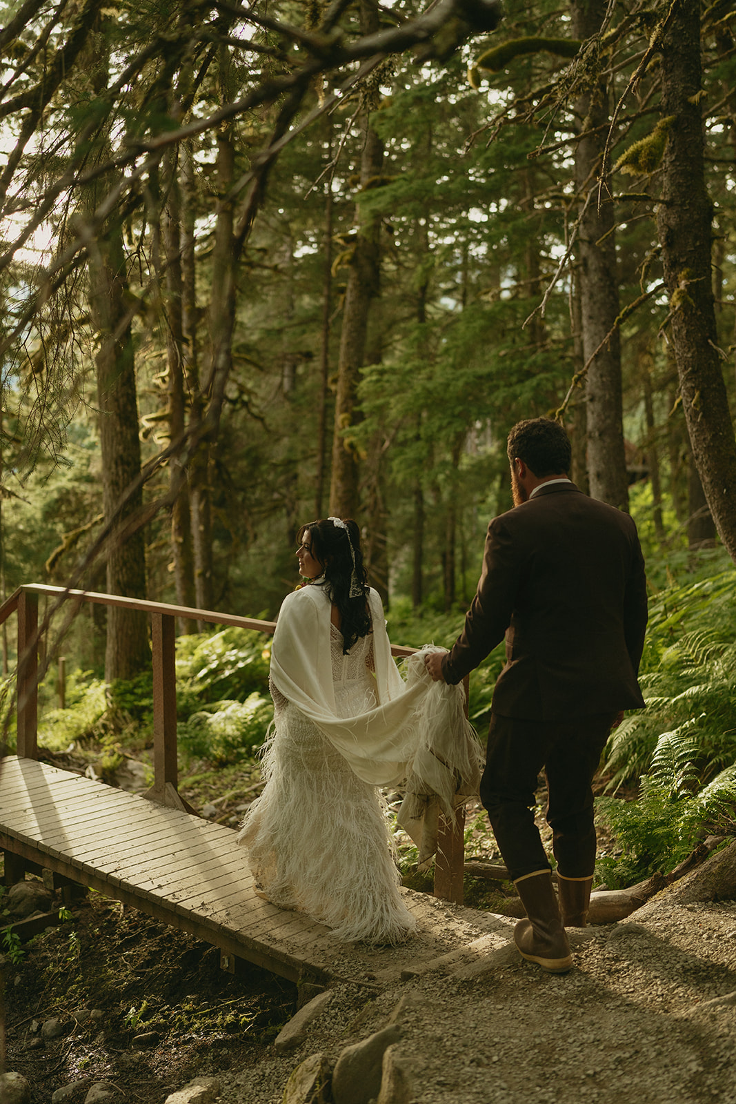 Groom holds bride's train while crossing wooden bridge in Alaska