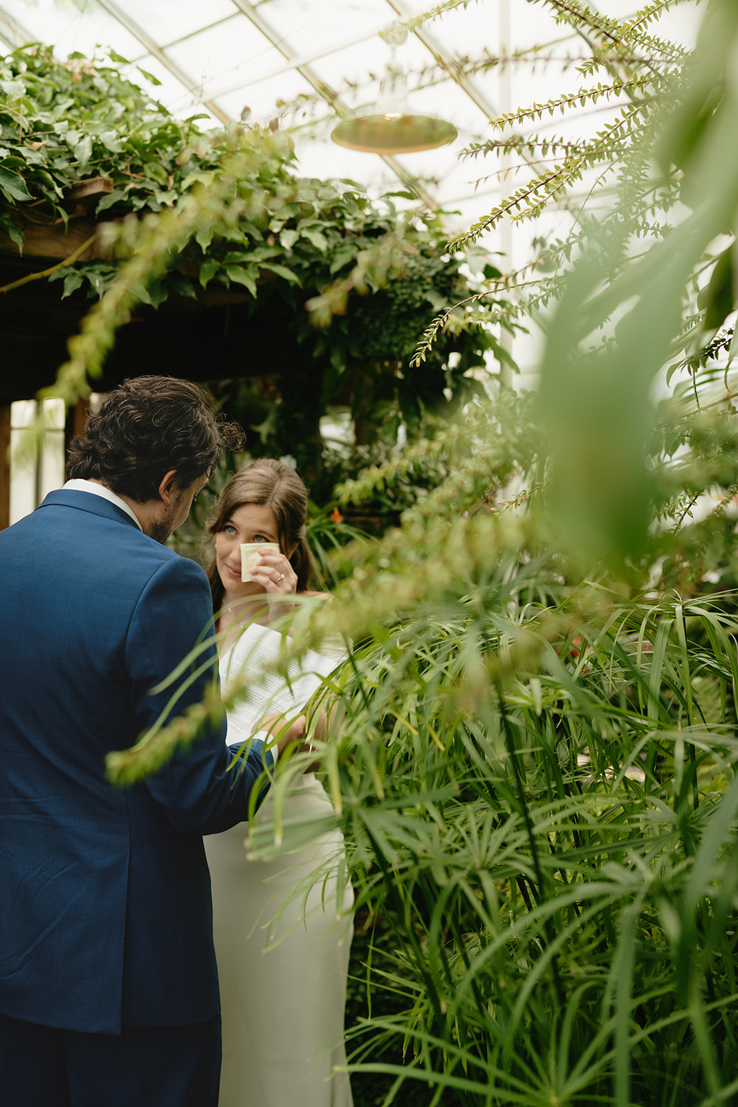 Bride wipes away a tear while sharing private vows with her partner, hidden among lush greenery at their greenhouse wedding.
