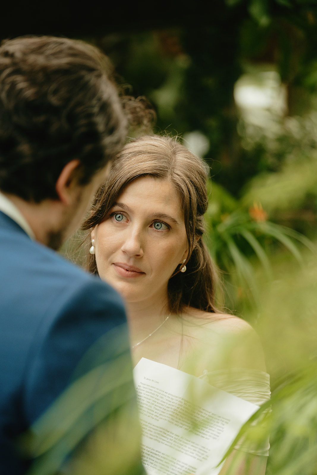 Bride looks up at her partner with emotion in her eyes while reading her vows, tucked into the foliage of their greenhouse wedding.