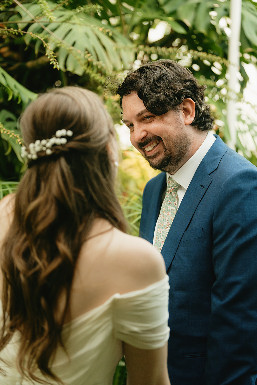 The groom gazes lovingly at his bride while exchanging vows, tropical plants filling the background of this greenhouse wedding.