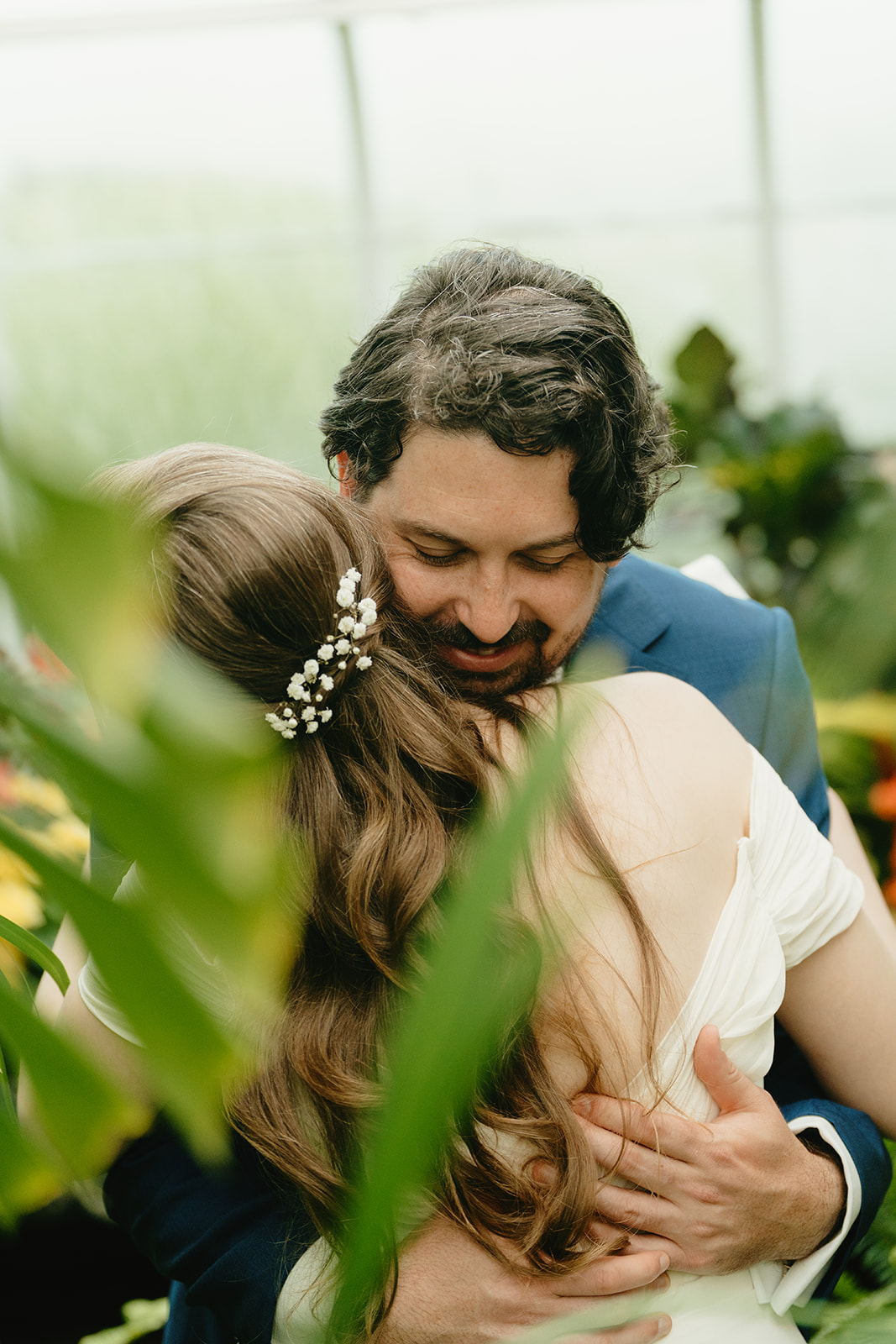 A groom embraces his bride with a peaceful smile, surrounded by the soft green tones of a lush greenhouse setting.