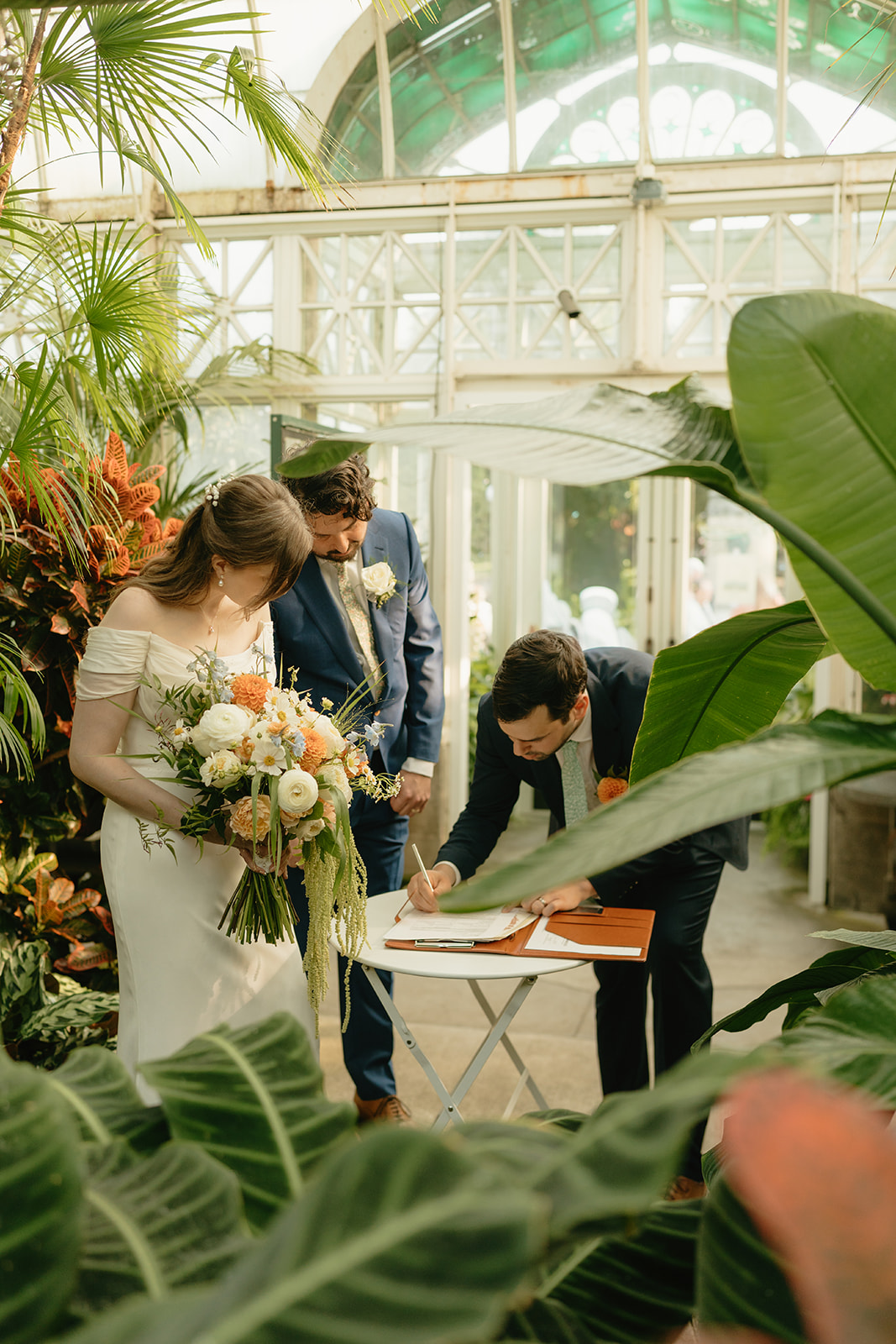 Couple signing their marriage license inside the greenhouse, surrounded by tropical plants and golden afternoon light.