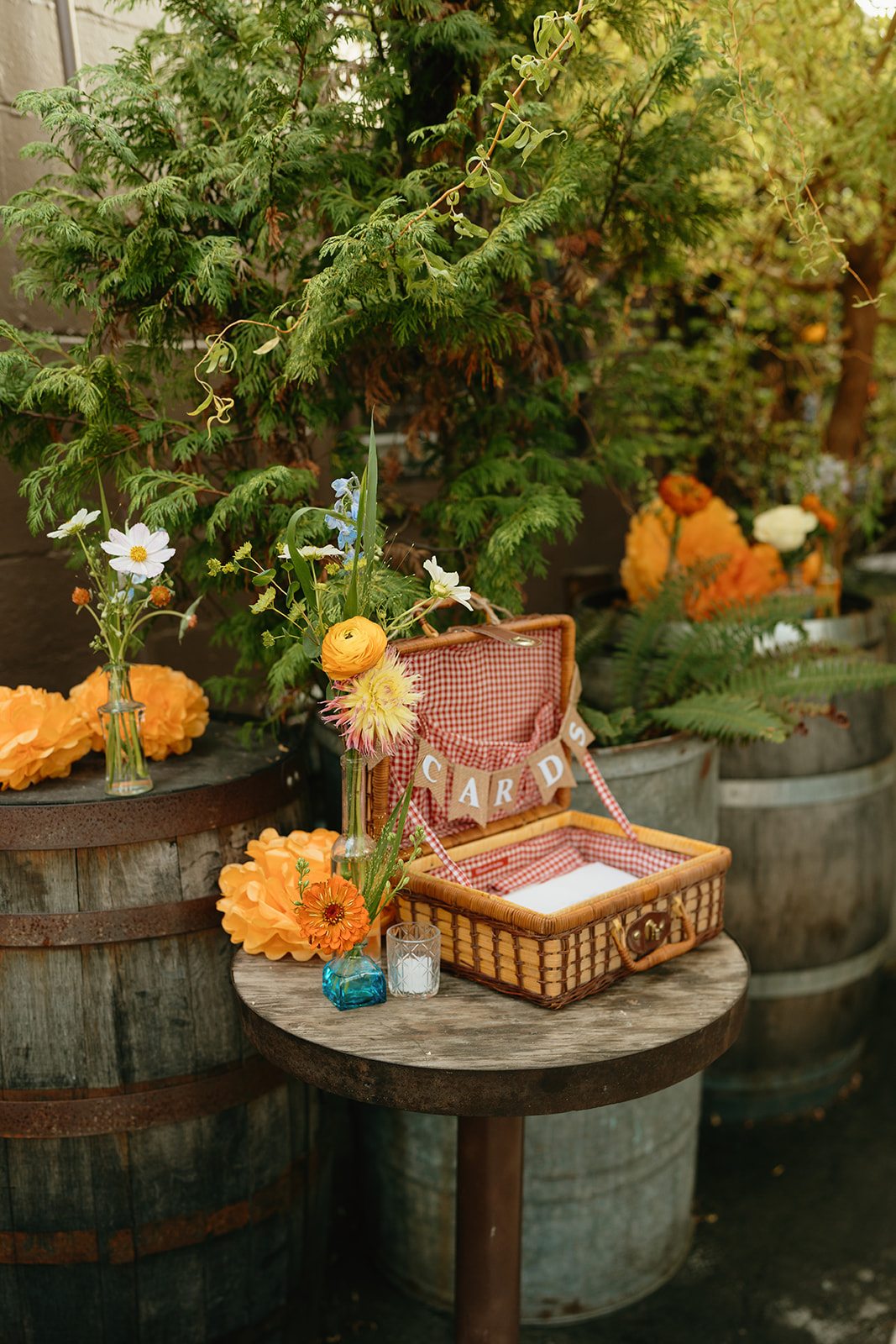 Charming card table display with wildflowers and a woven basket, nestled among barrels and greenery for a whimsical wedding touch.