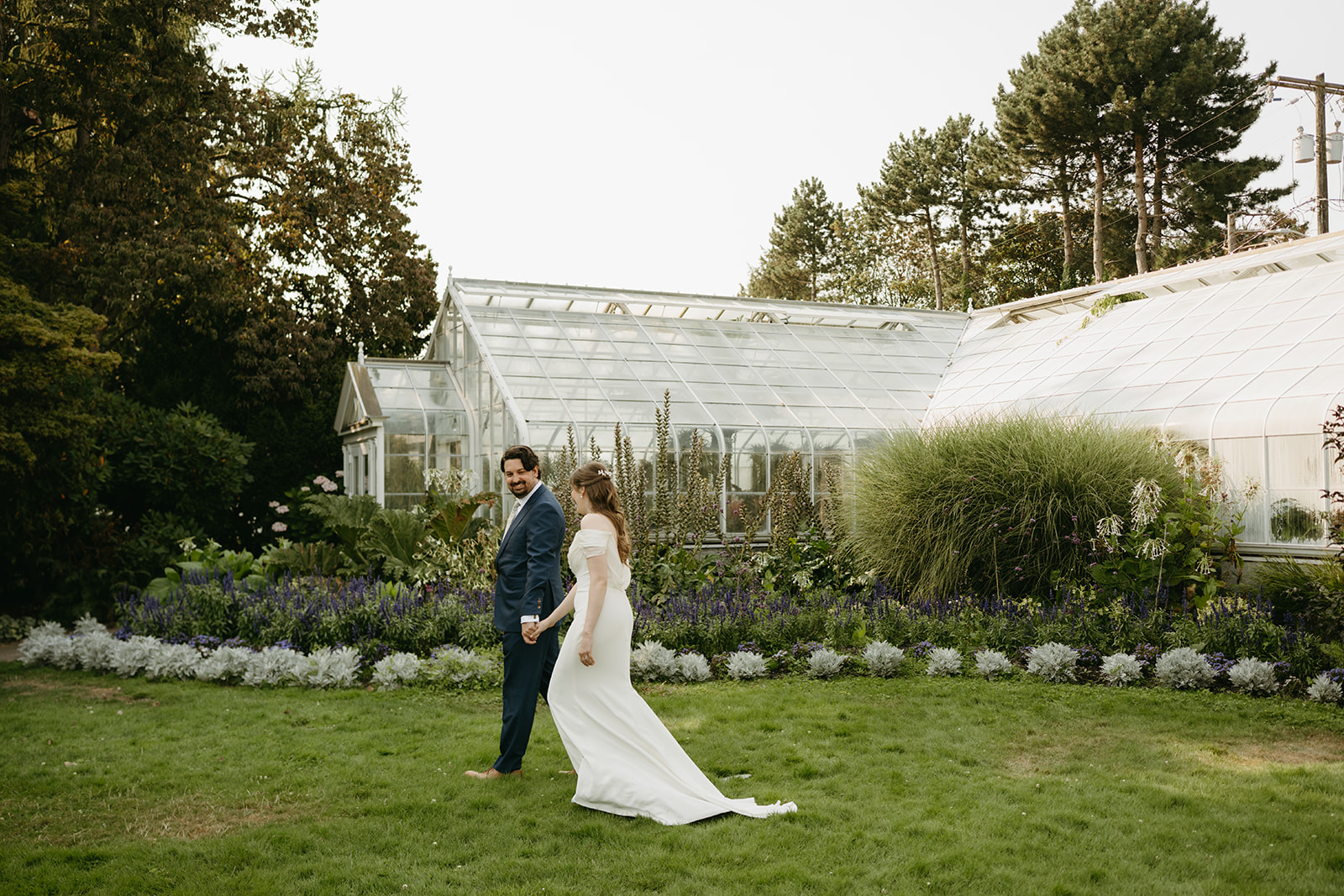 Couple walks hand in hand outside a historic glass conservatory, framed by flowering gardens after their greenhouse wedding.