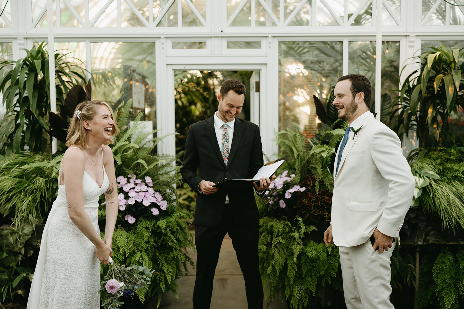 A joyful bride and groom laughing with their officiant during a greenhouse wedding ceremony, surrounded by vibrant ferns and tropical plants.