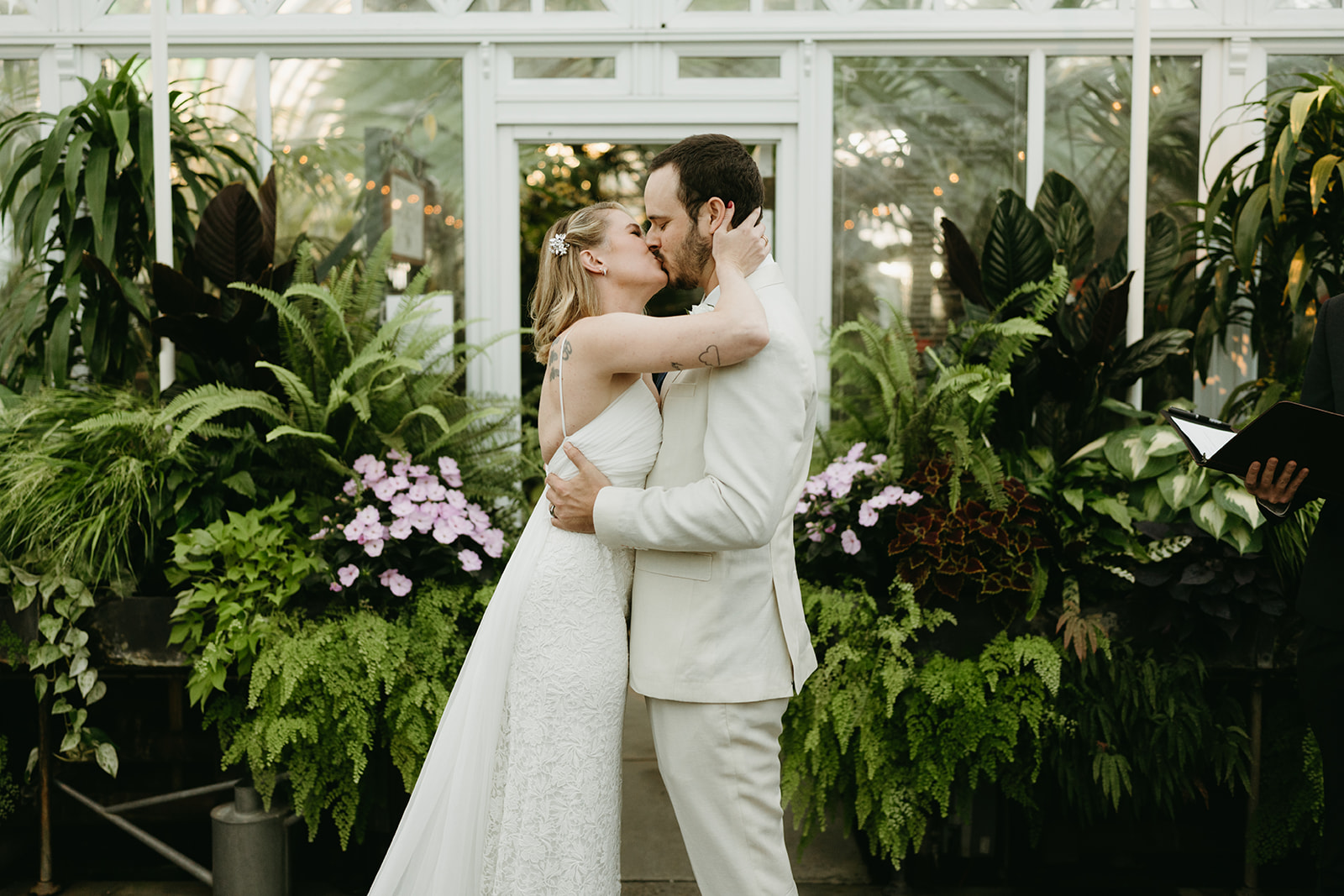 A just-married couple shares a kiss in front of lush tropical plants and soft florals during their intimate greenhouse wedding ceremony.