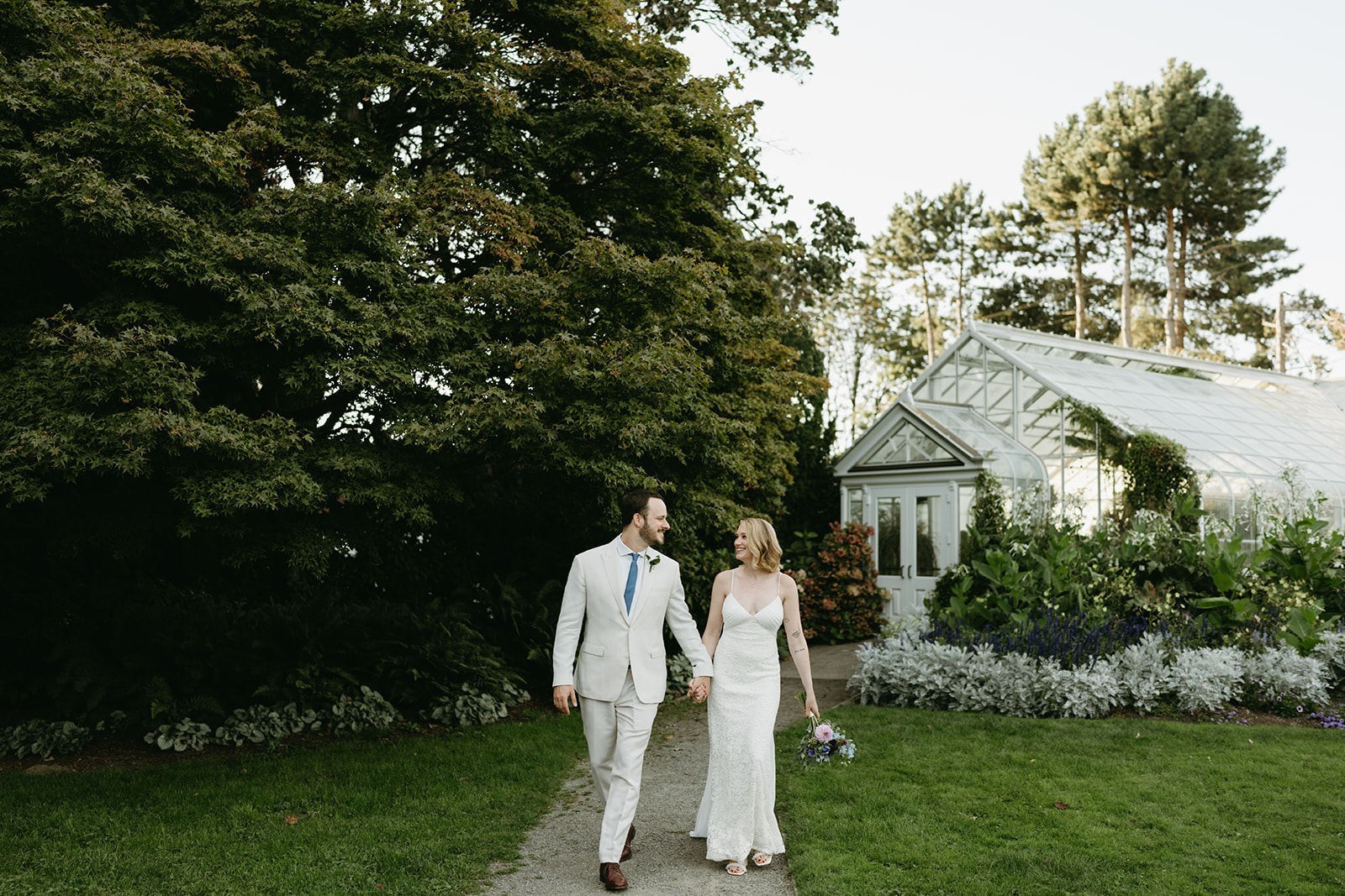 Bride and groom walking hand in hand outside the conservatory, smiling at each other after their joyful greenhouse wedding ceremony.