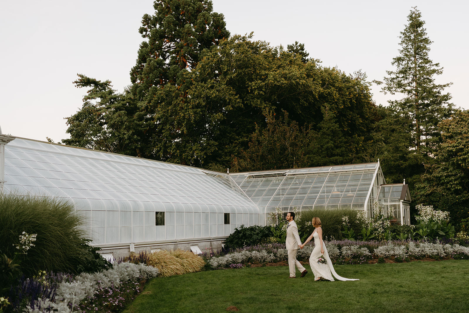 Couple walking hand in hand outside a historic greenhouse, surrounded by lush gardens and tall trees during golden hour.
