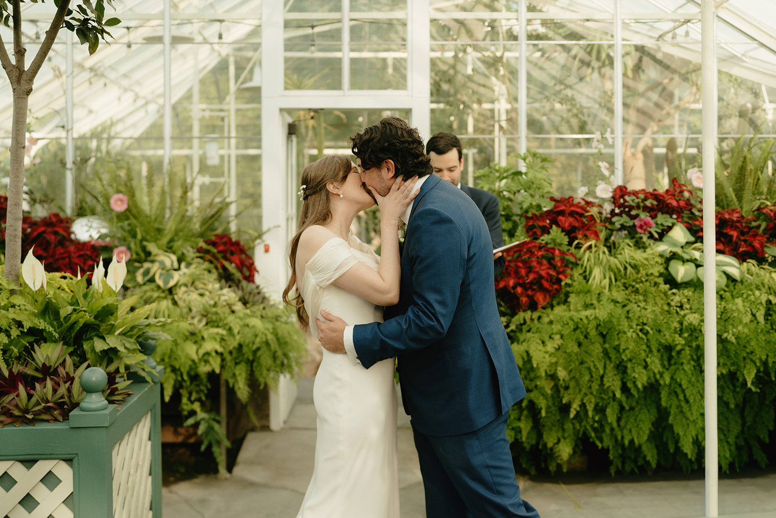 Bride and groom share their first kiss in a lush greenhouse wedding ceremony, surrounded by bold greenery and red blooms.