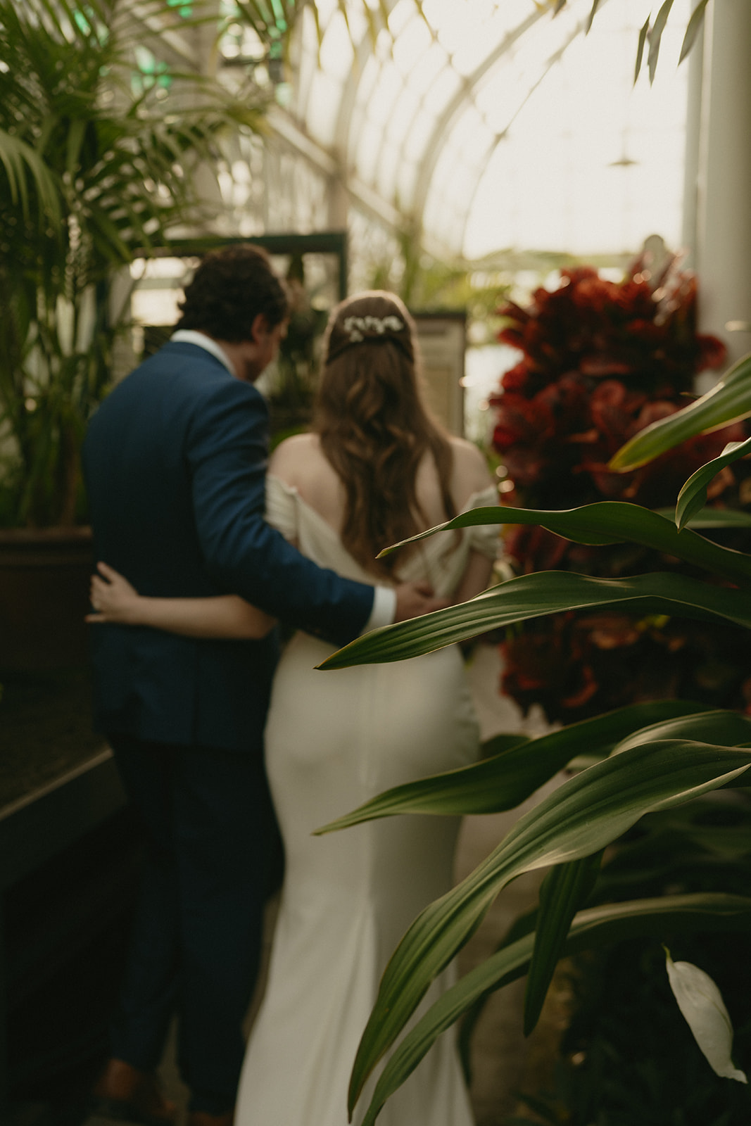 Newlyweds walk arm-in-arm through a greenhouse wedding space, framed by tropical plants and warm natural light.