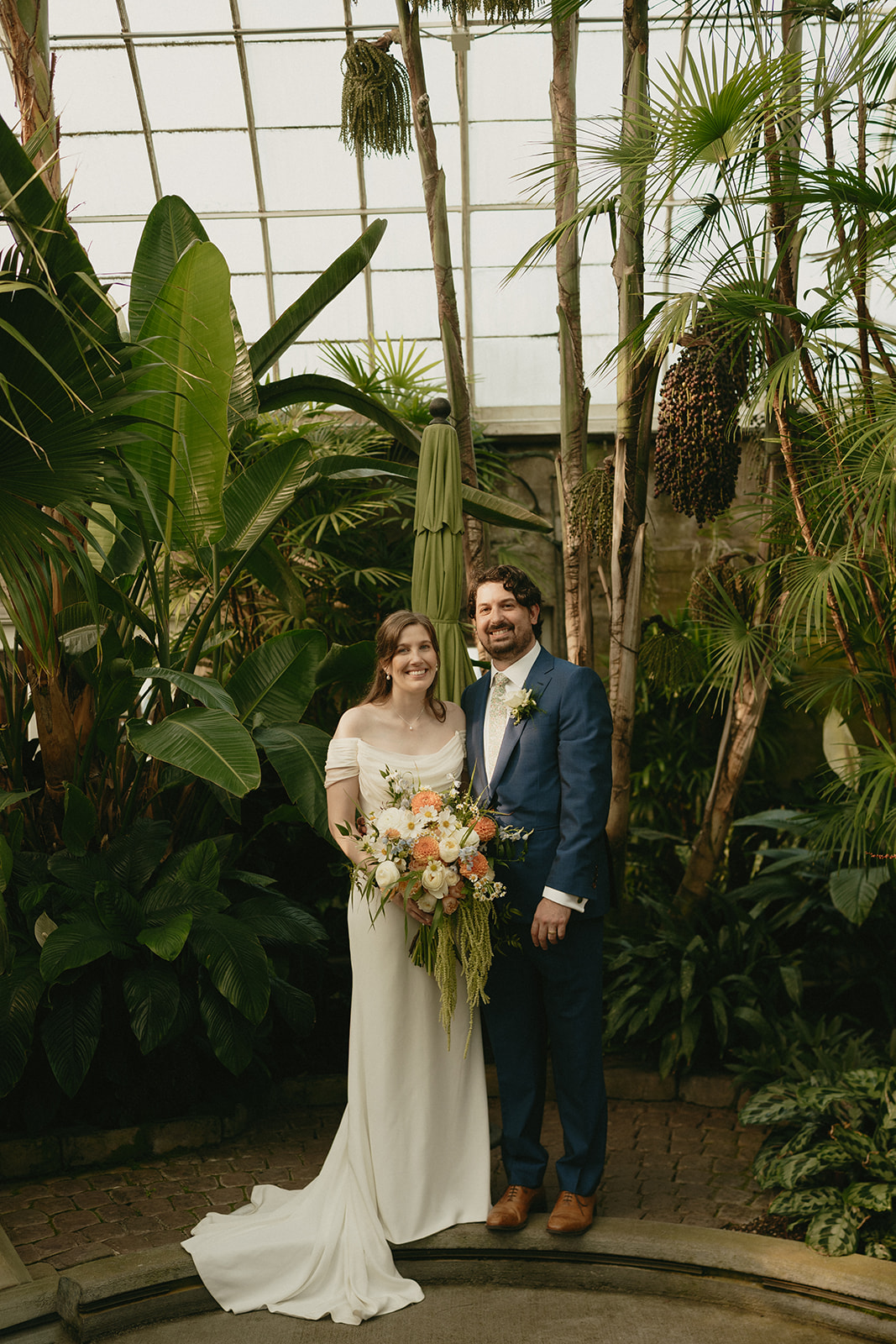 Bride and groom standing together in front of tropical plants, smiling warmly after their intimate greenhouse wedding.