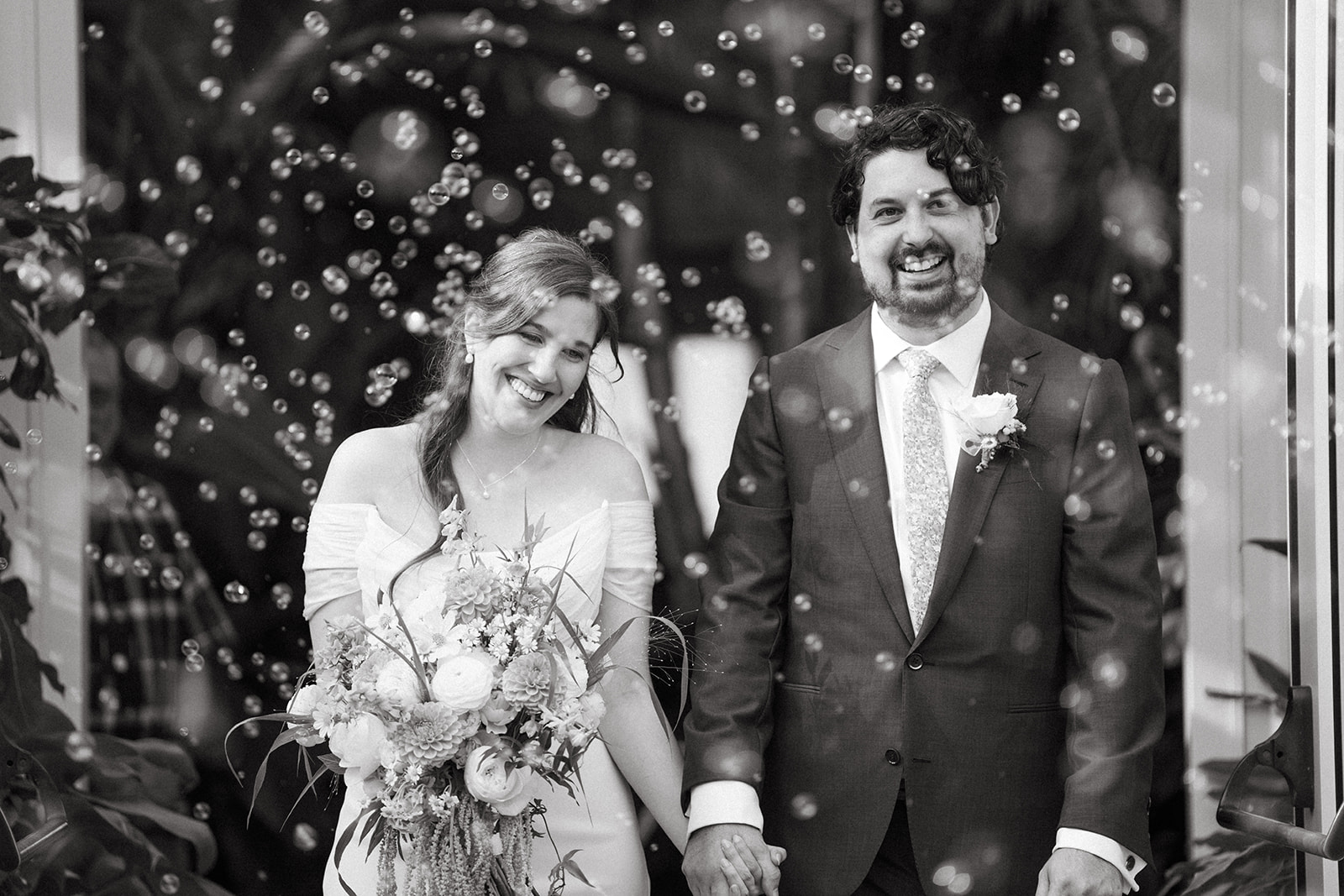 Newlyweds walk through a bubble exit with joy and laughter, captured in dreamy black and white outside the greenhouse.