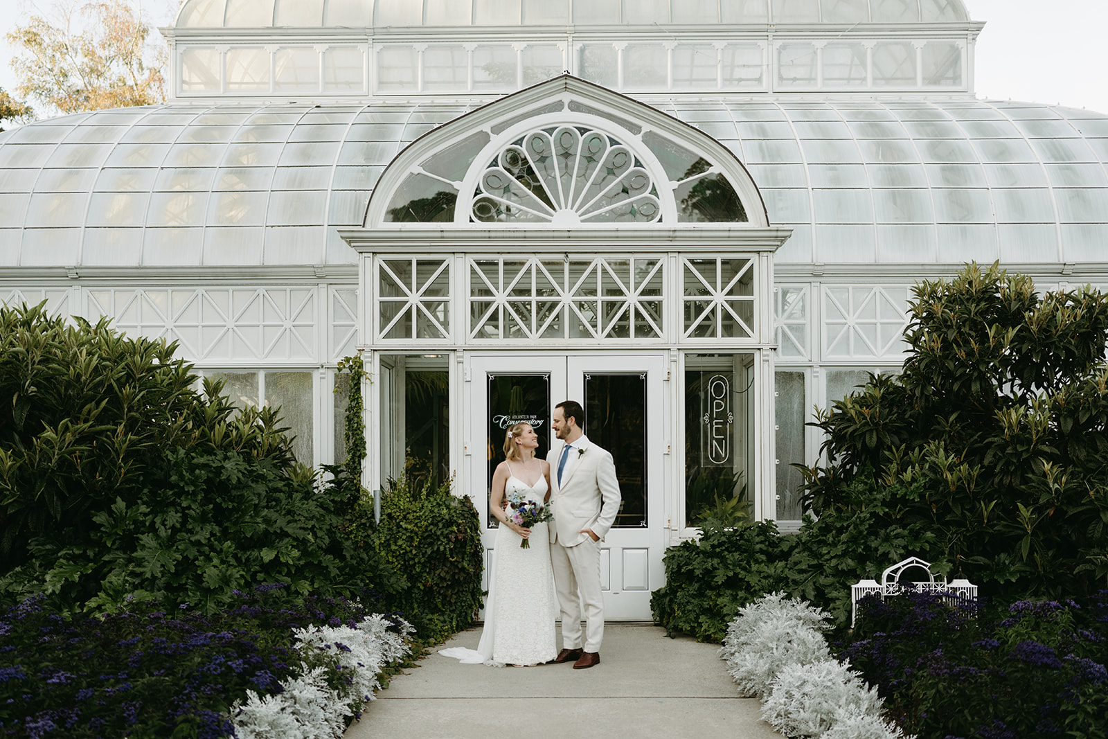 Couple standing in front of a vintage glasshouse entrance, looking at each other with joy and love after their greenhouse wedding.