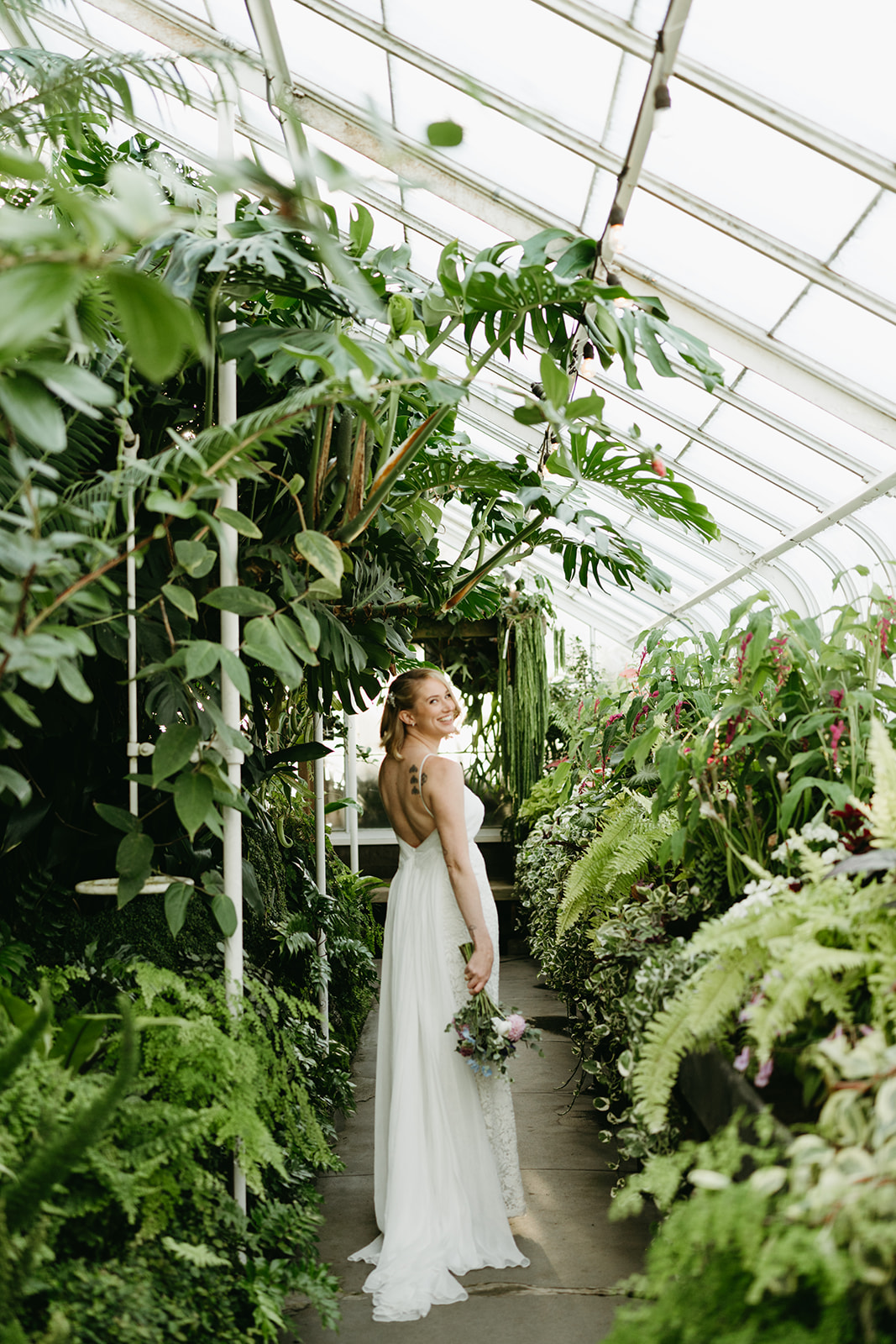 Bride holding her bouquet and smiling over her shoulder in a corridor of greenery during a serene greenhouse wedding portrait session.