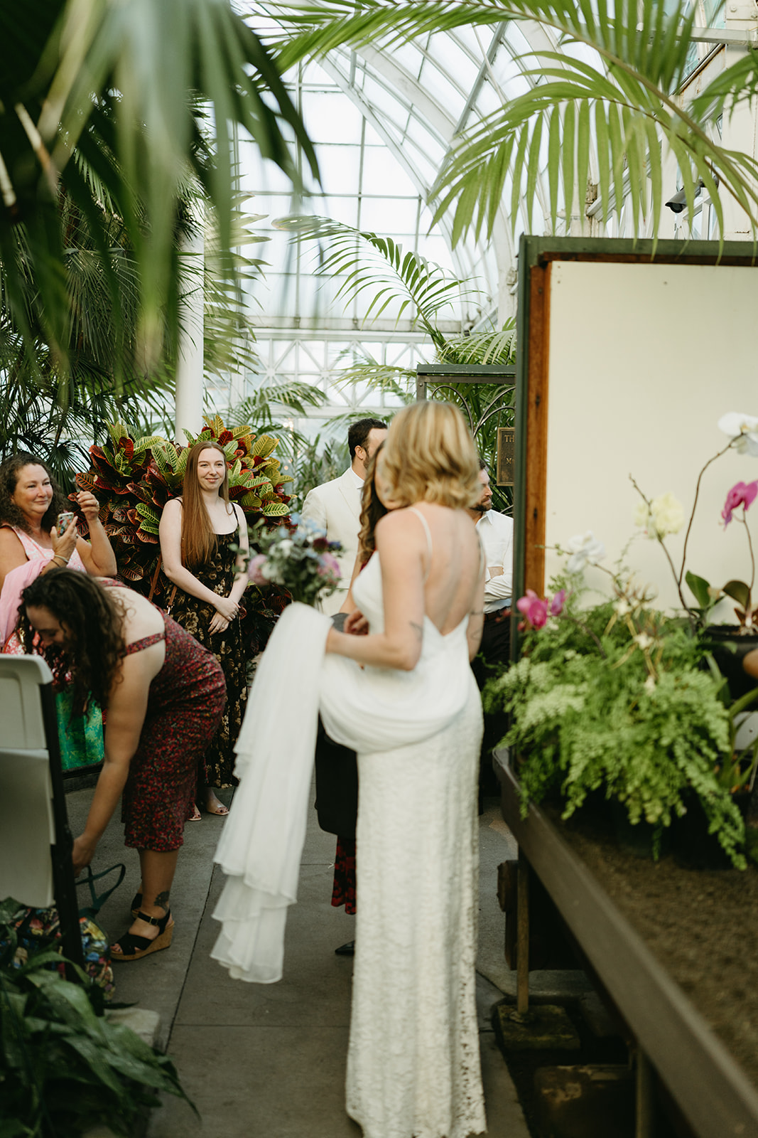 Bride mingling with guests inside the greenhouse, holding her dress and bouquet as celebration unfolds around her.
