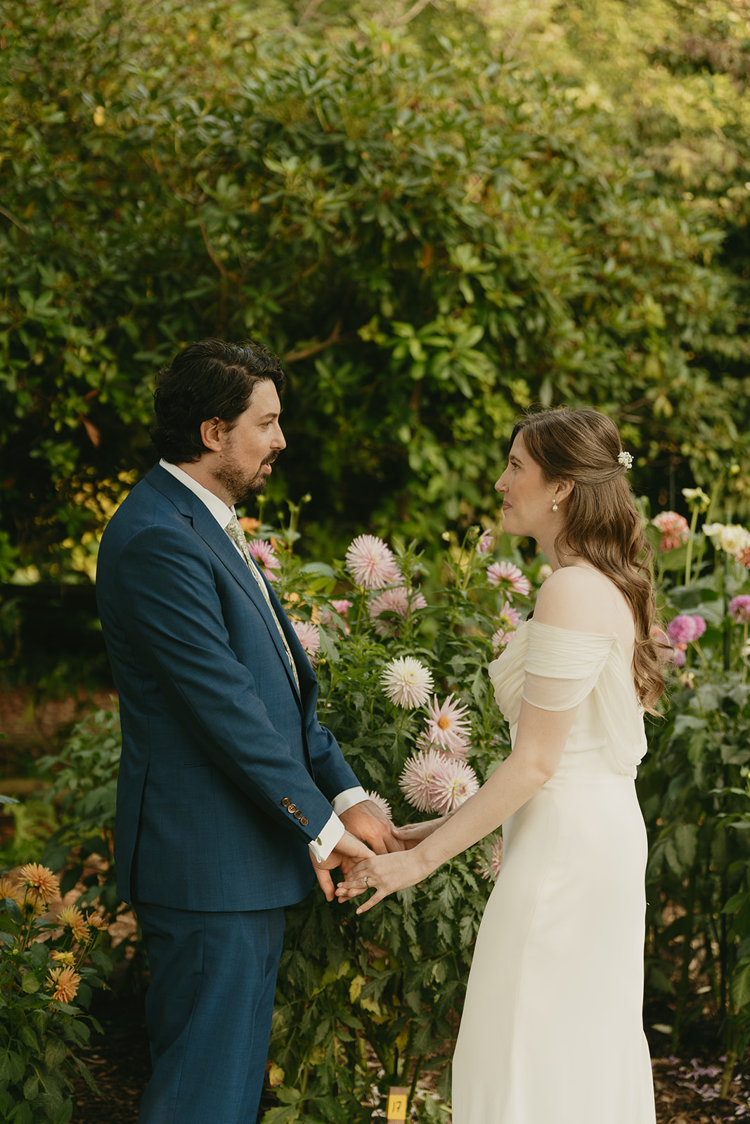 Bride and groom standing in a garden full of blooming dahlias, holding hands and soaking in the moment together.