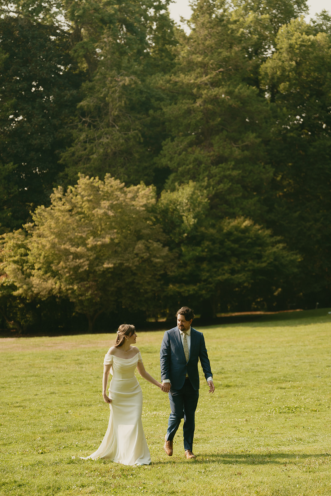Bride in an off-the-shoulder gown walks through an open field with her groom beneath tall summer trees.