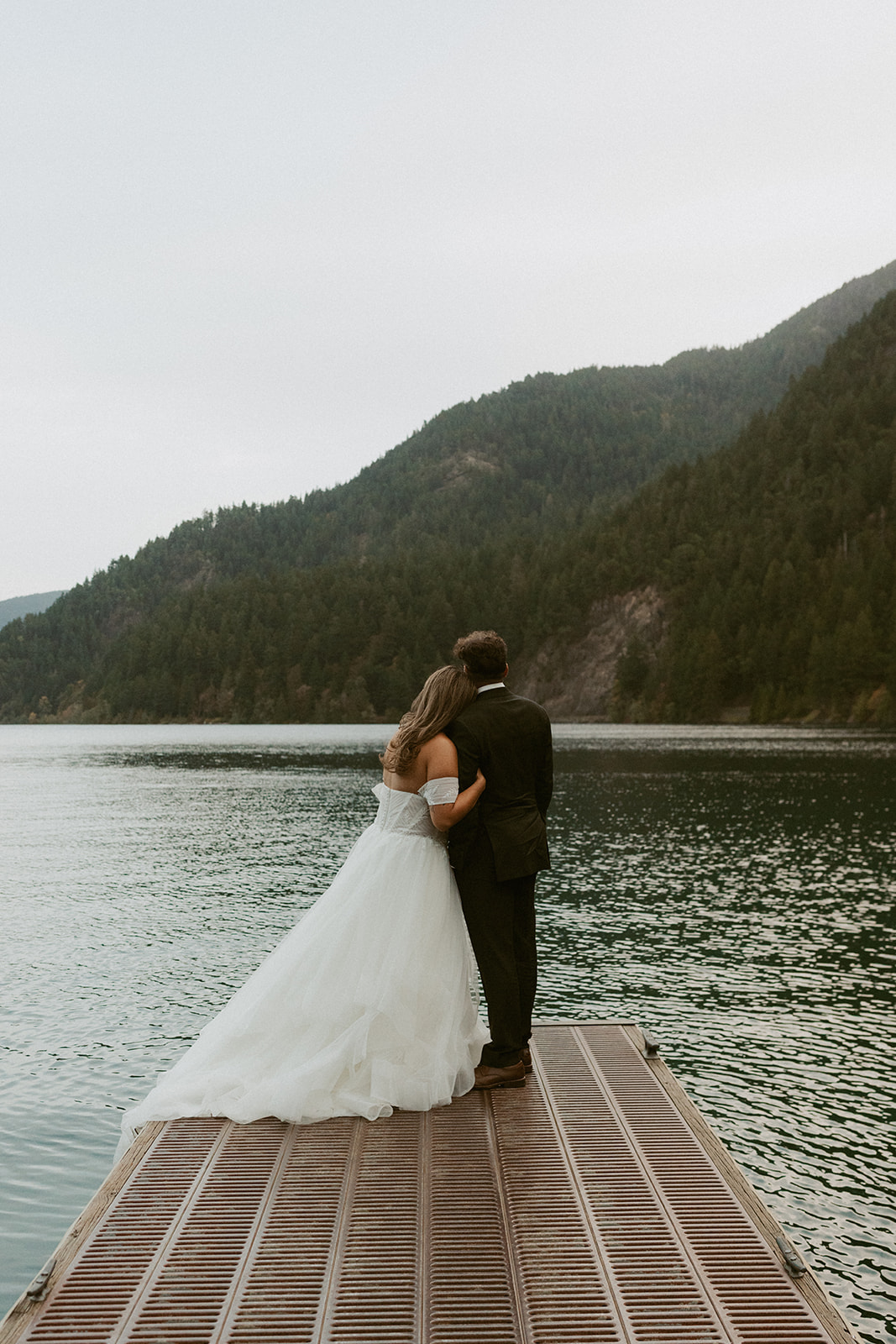 The couple stands together at the end of a wooden dock, overlooking the still waters of Lake Crescent during their Olympic National Park elopement.