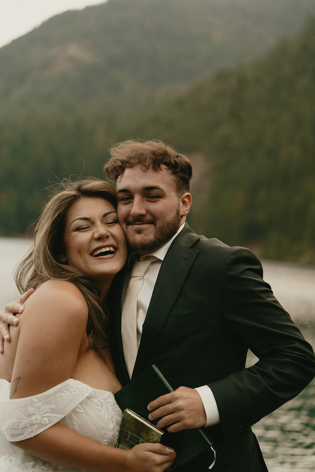 A joyful couple embraces after exchanging vows, laughing together with a serene mountain lake behind them.