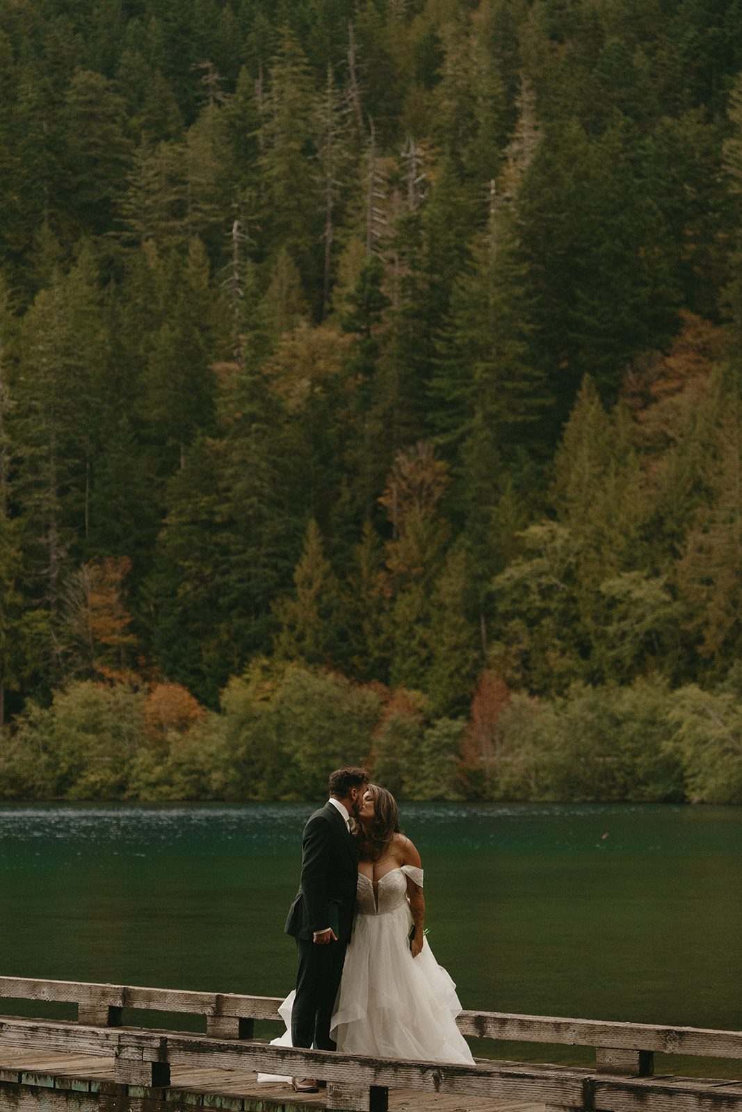 The bride and groom share a kiss on a dock above emerald green water, embraced by the quiet stillness of the Olympic National Park elopement landscape.