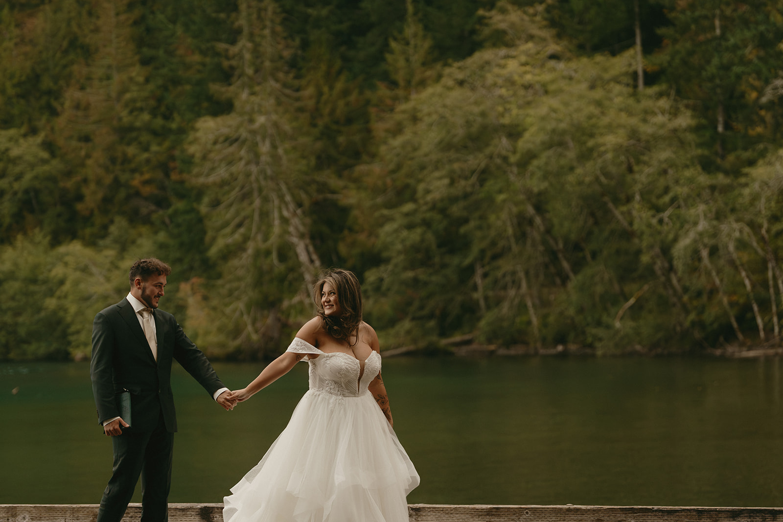 The couple holds hands and smiles as they walk along the edge of a still lake, the water reflecting the evergreen forest around them.